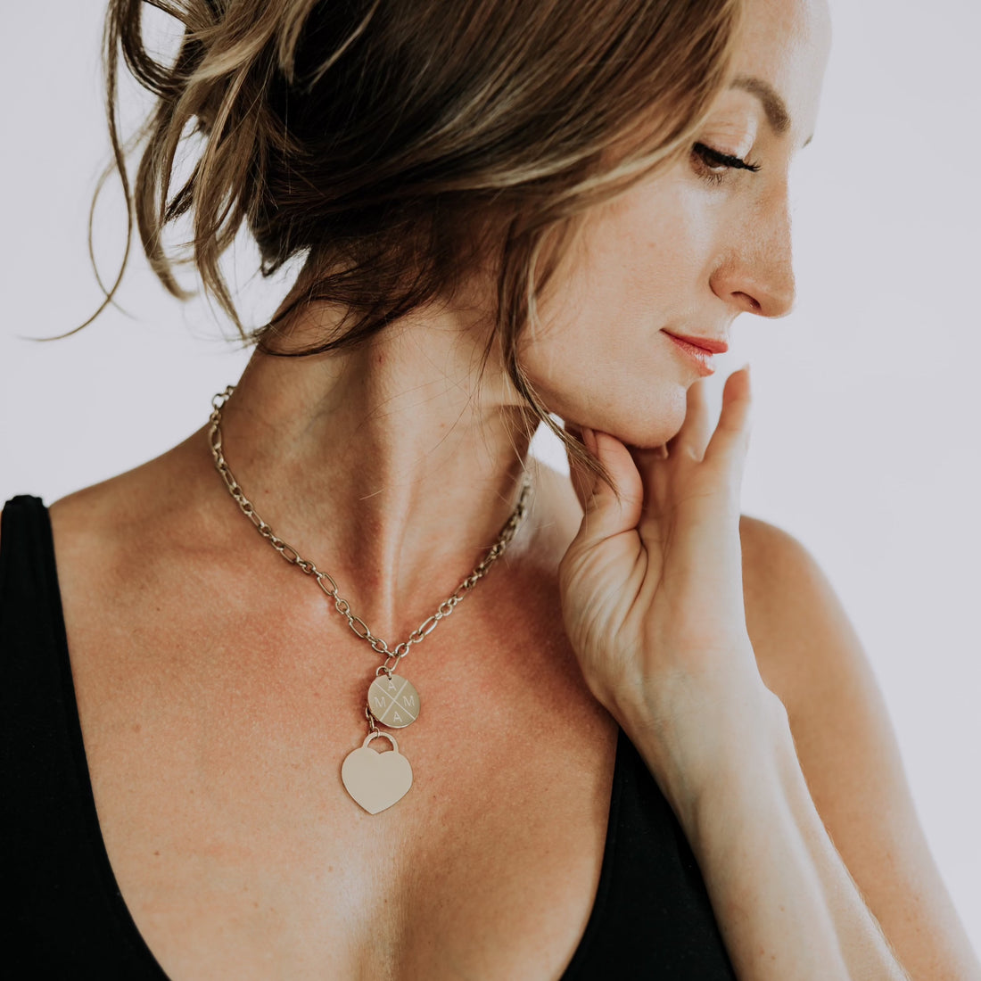 A woman with light brown hair pulled back gazes downwards with her eyes closed, resting her hand on her neck. She wears a Mama X™ Brand gold stainless steel necklace featuring two pendants: a round disk and a heart shape from the Mama X™ Gold Layered Necklace collection. She is dressed in a black sleeveless top against a neutral background.