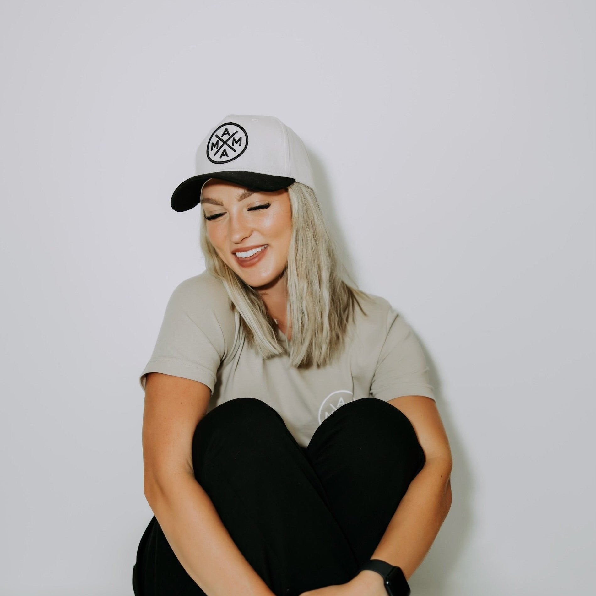 A woman with long blonde hair sits against a white wall, smiling with her eyes closed. She wears a cream-colored baseball style cap from Mama X™ and has on a light gray T-shirt and black pants, with a smartwatch on her wrist. The setting is calm and minimalistic.