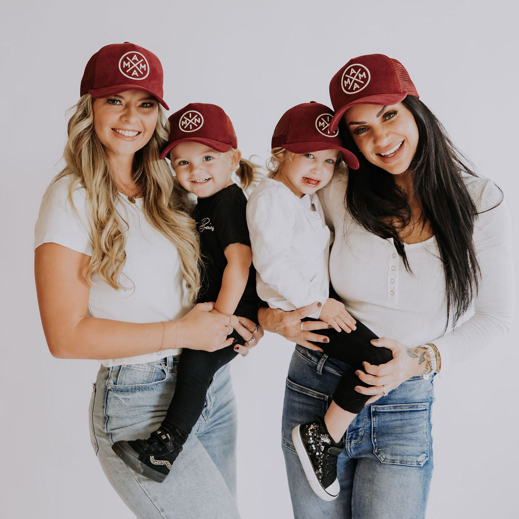 Two women and two girls pose together, smiling in matching Mama X™ Brand Mom &amp; Mini Suede Bundle hats with a white logo. The women wear jeans and tops, while the girls are in black and white outfits against a white background.