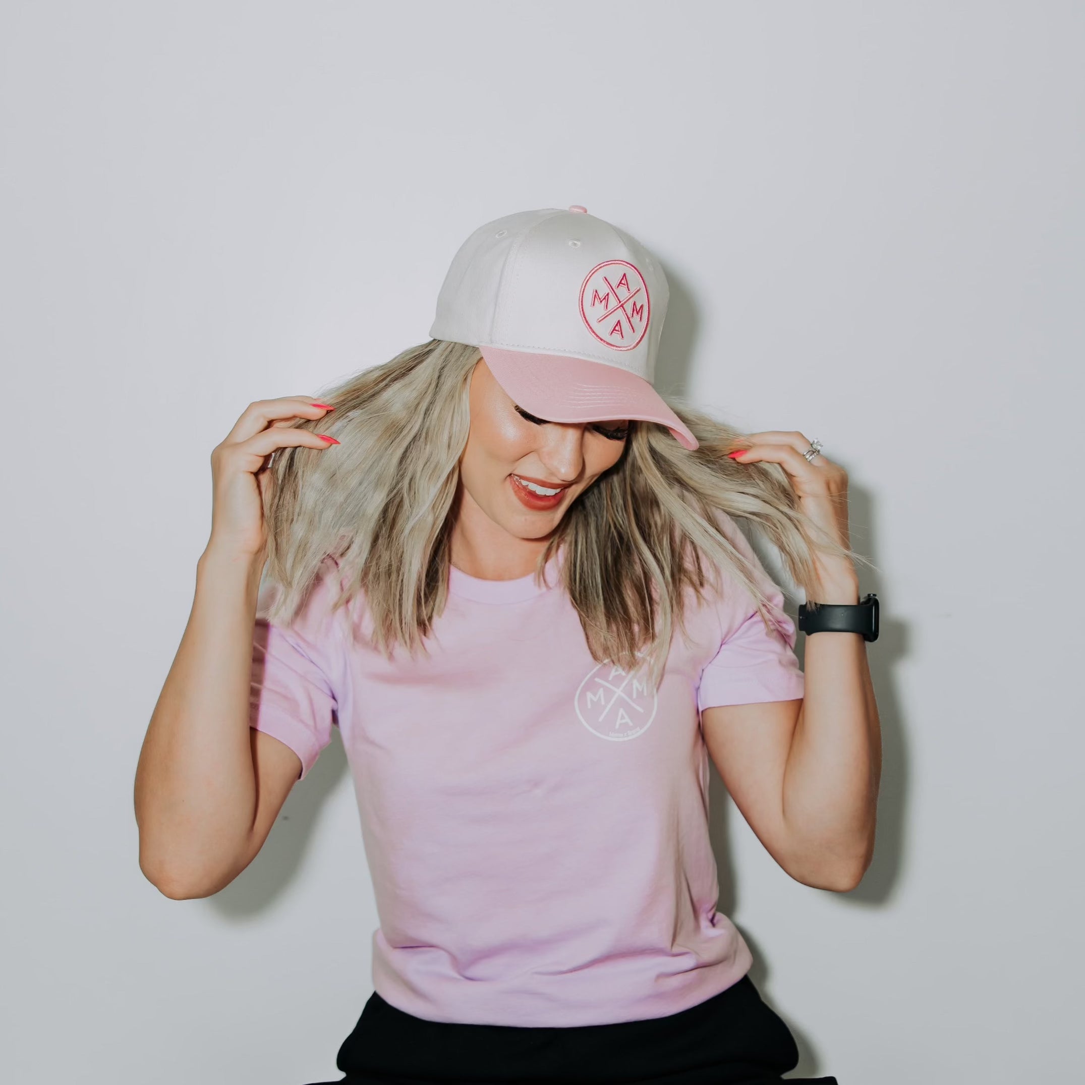 A person with long, light blonde hair wears a pink and white MAMAM hat, a lilac Mama X™ T-Shirt - Lilac in unisex sizing from the Mama X™ Brand, and a black smartwatch. They are smiling, looking down, and gently holding their hair with both hands against a plain white background.