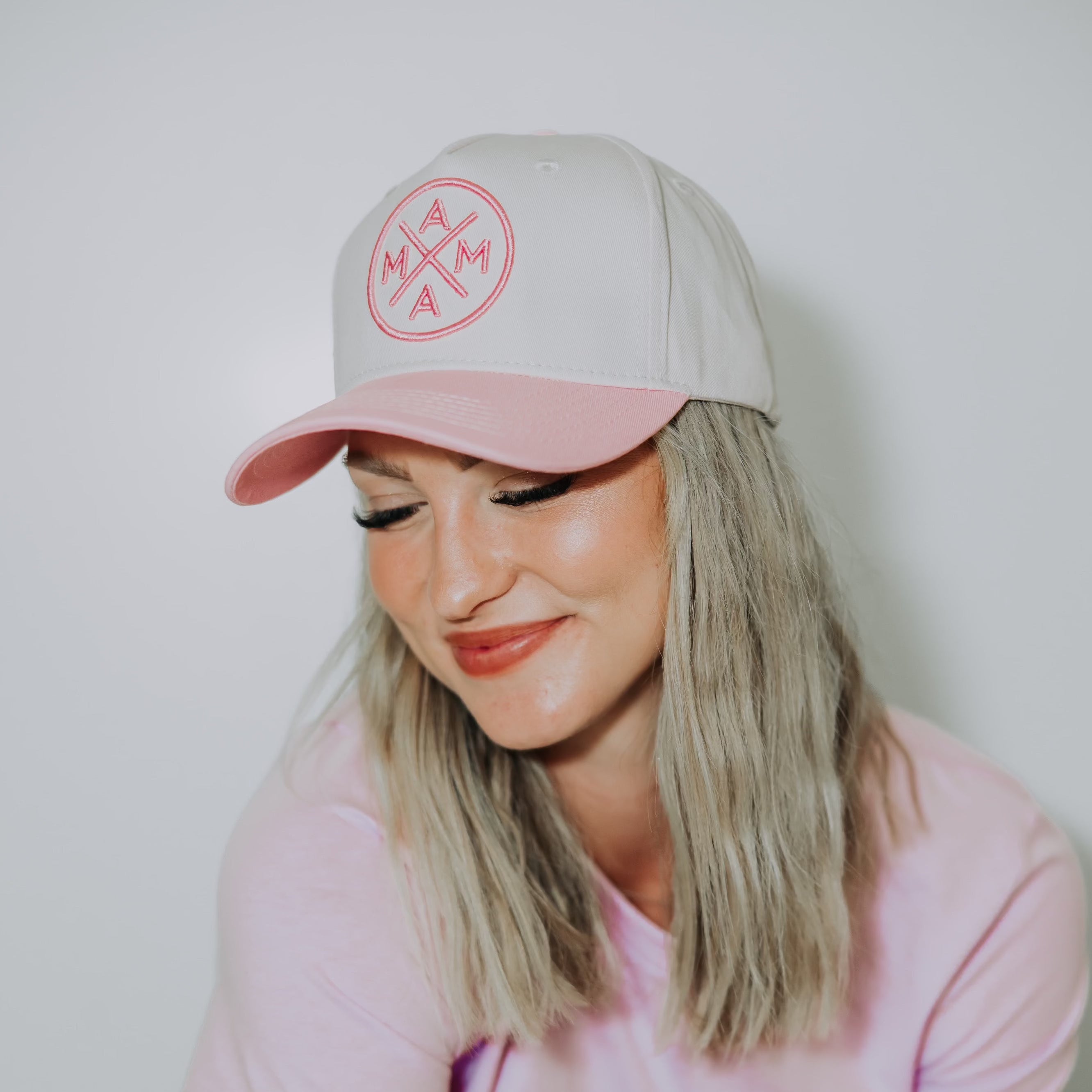 A woman with light blonde hair is wearing a light pink and white baseball cap featuring the 