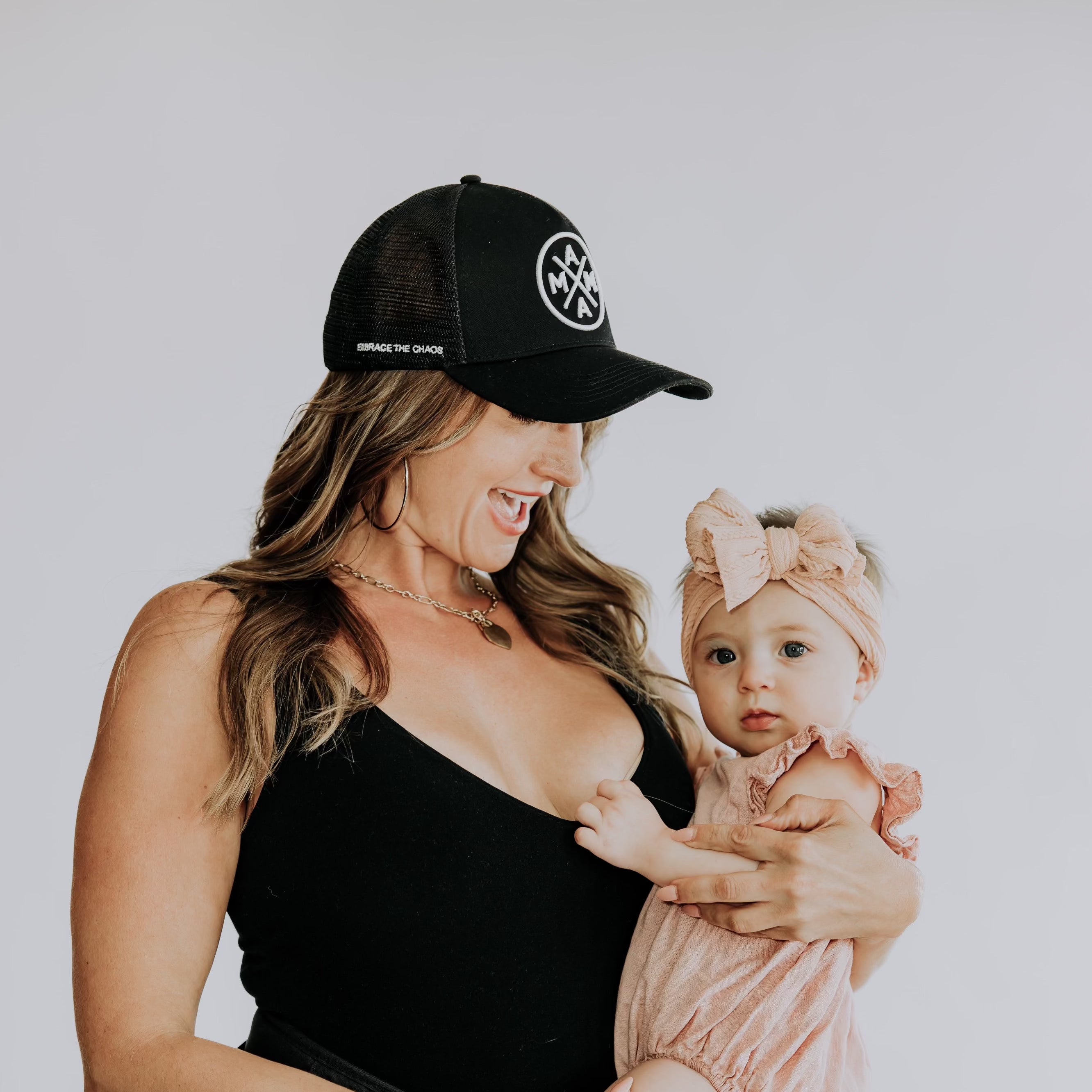 A woman in a black tank top and a chic Mama X™ Embrace The Chaos Trucker Hat by Mama X™ Brand holds a baby in a pink outfit and headband. The woman smiles joyfully at the baby, who looks at the camera with a neutral expression. The background is plain white.