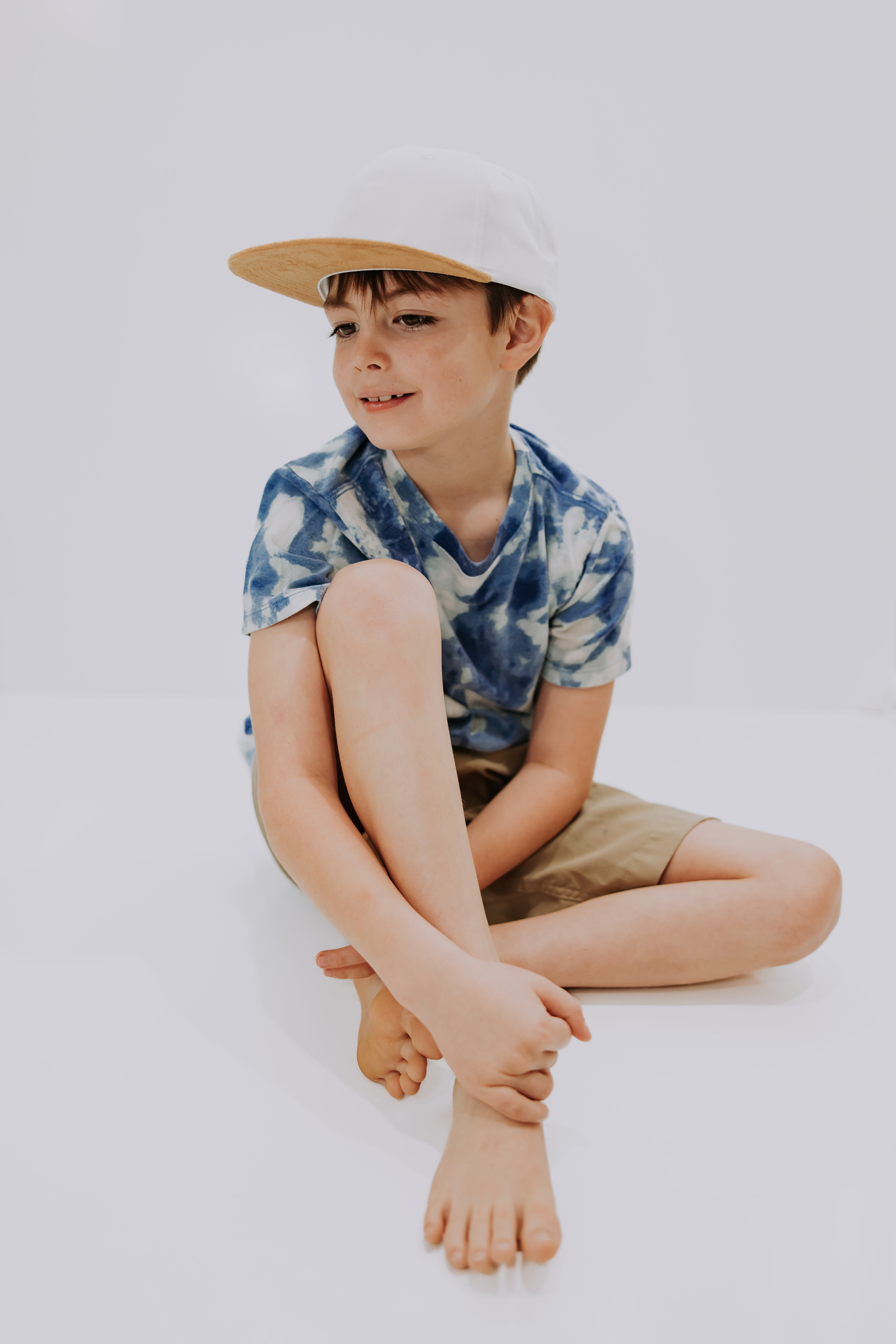 A young boy sits on the floor with his legs crossed, wearing a blue tie-dye t-shirt, beige shorts, and a stylish Mama X™ Brand White With Brown Suede Bill Trucker hat. He is smiling gently and looking off to the side against a plain white background.