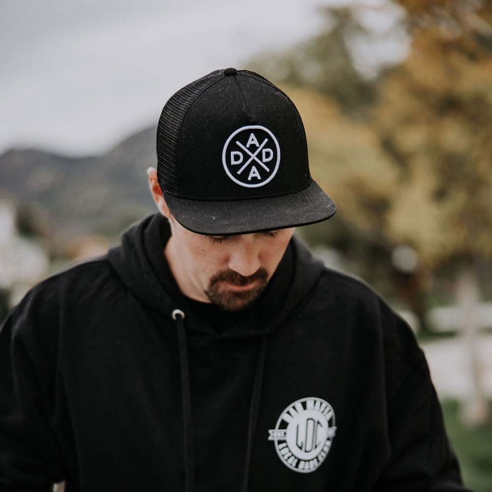 A man wearing a black Dada X™ Premium Trucker Hat with a stitched 3D logo from Mama X™ Brand looks down. He is dressed in a black hoodie featuring a white circular logo on the chest. The background is blurred, showcasing green and yellow foliage along with mountains.