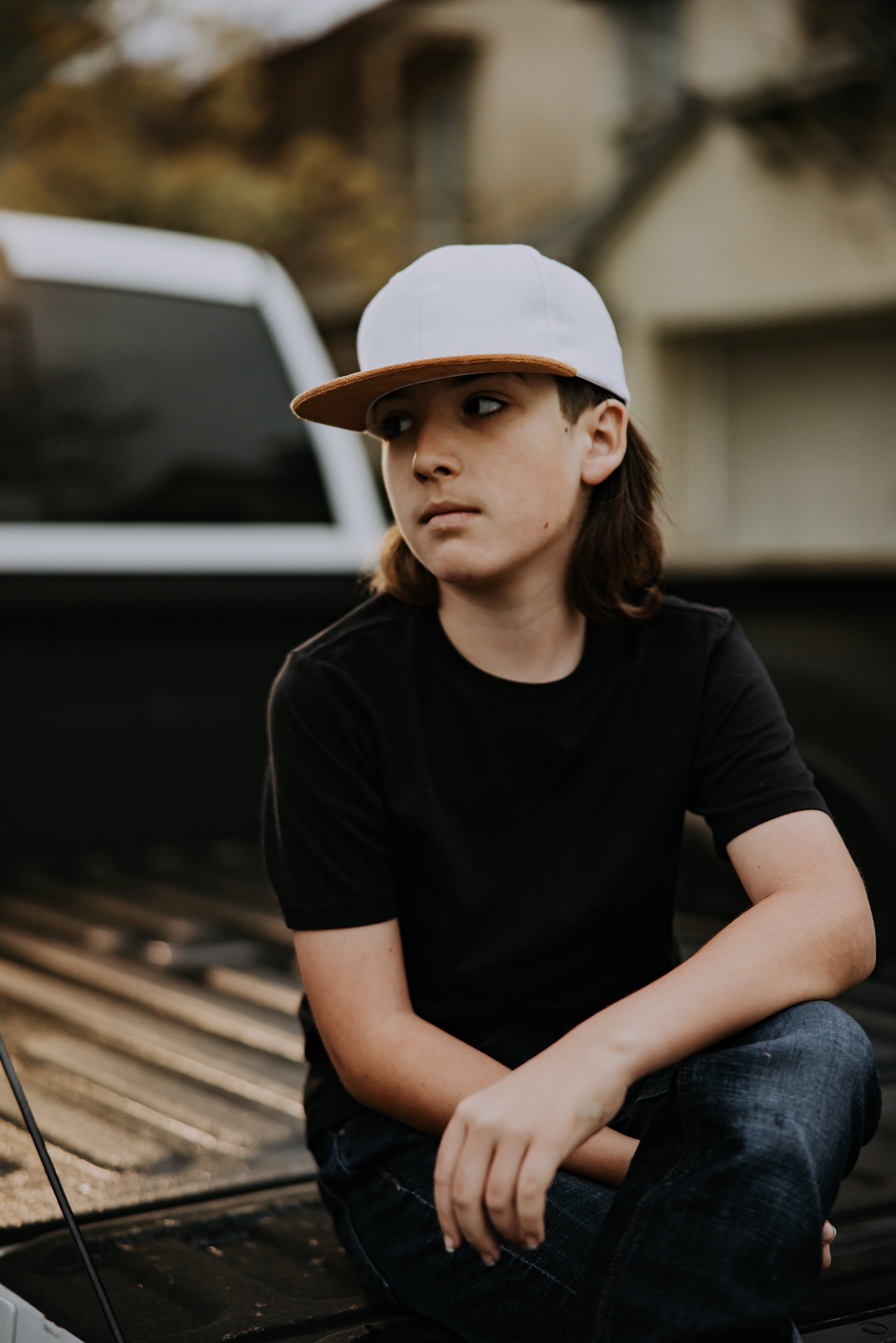 A young person with long hair is sitting in the bed of a pickup truck, wearing a Mama X™ Brand White With Brown Suede Bill Trucker and a black t-shirt. They have a pensive expression, looking to the side with soft lighting highlighting their face. The background is slightly blurred, showing part of the truck and its surroundings.