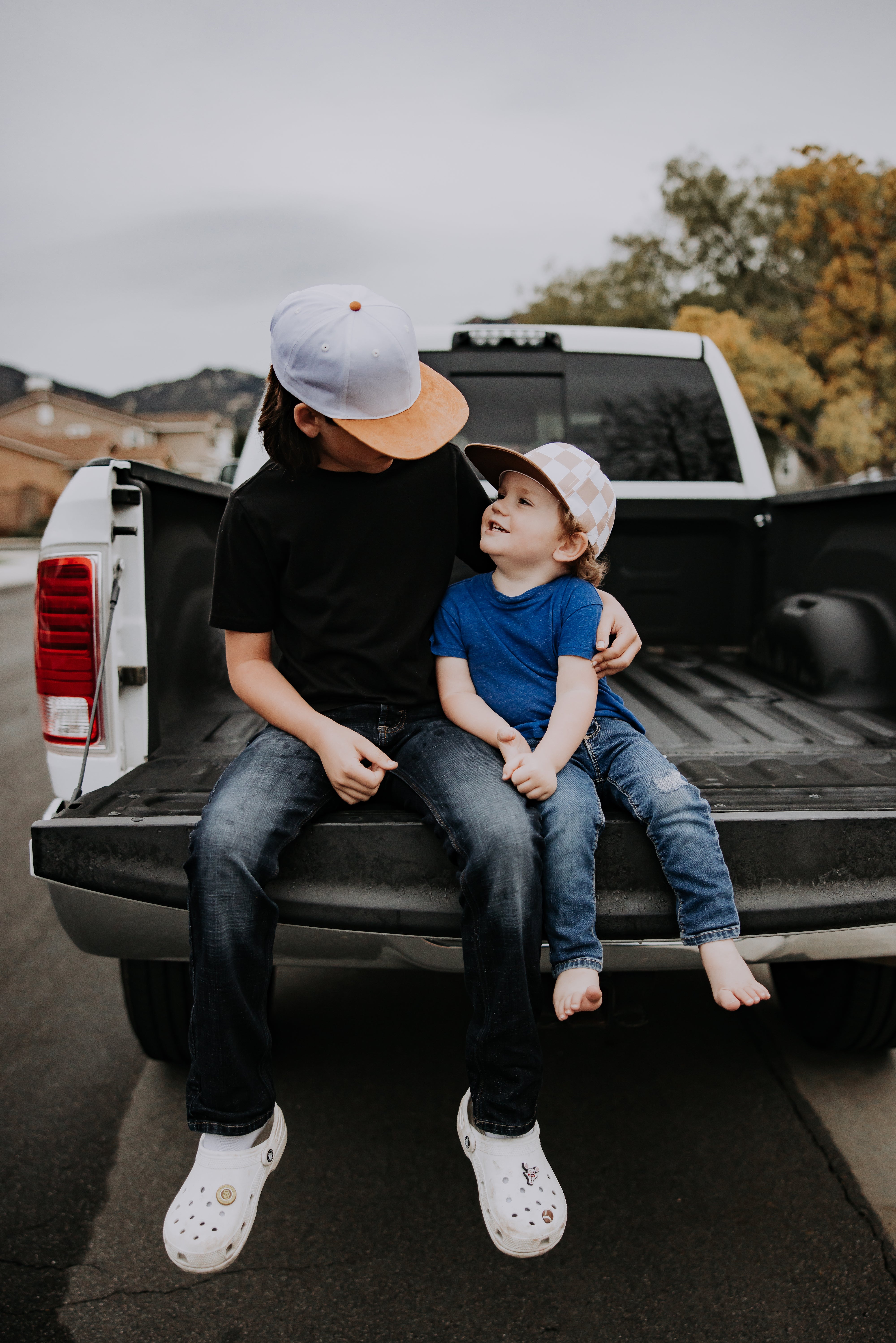 Two young children sit on the open tailgate of a pickup truck. The older child, in a black shirt and jeans with a **Brown And White Checkered Trucker** cap from *Mama X™ Brand,* sits beside the younger one. The toddler, dressed in a blue shirt and jeans, looks up smiling. They appear to be enjoying a moment together outdoors.