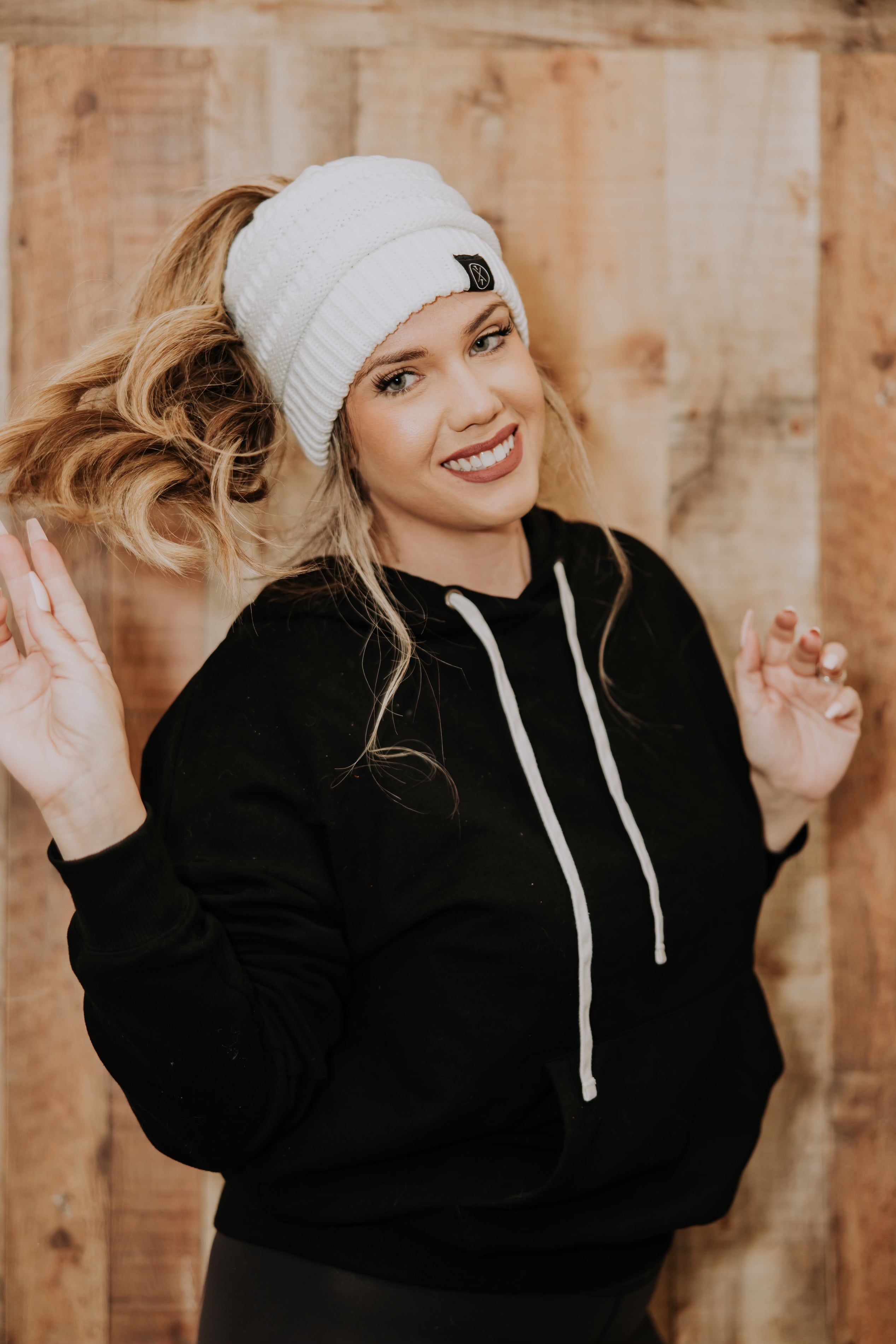 In front of a wooden wall, a smiling person with long wavy blonde hair poses while wearing Mama X™ Brand's Knit Beanie For Adults With Ponytail Hole, which allows their ponytail to stick out. They are also dressed in a black hoodie and have their hands raised near their shoulders, exuding a playful and cheerful vibe.