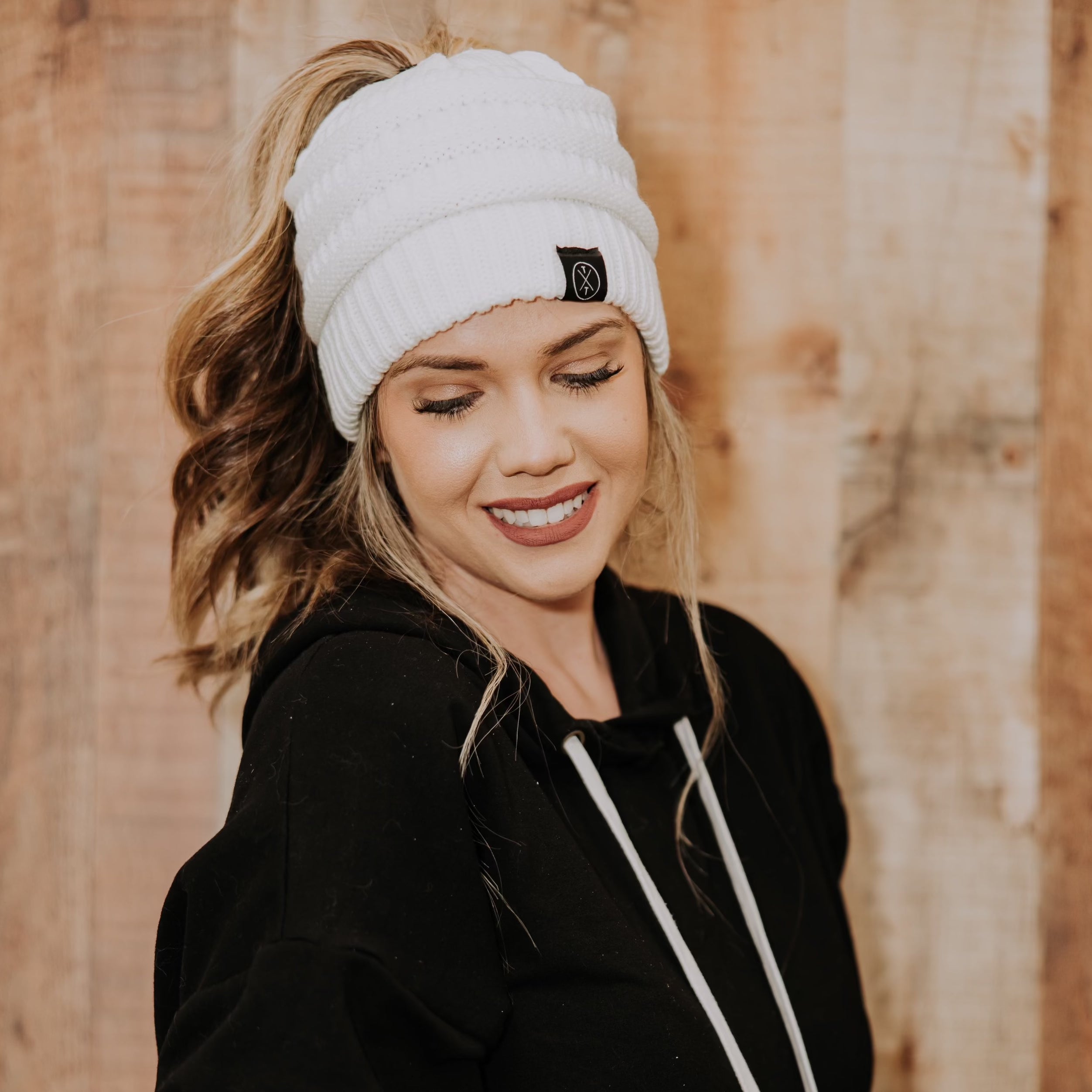 A young woman wearing a Mama X™ Brand Knit Beanie For Adults With Ponytail Hole and a black hoodie with white drawstrings smiles and looks down. Her wavy brown hair peeks out from beneath the beanie as she stands in front of a light wooden wall.