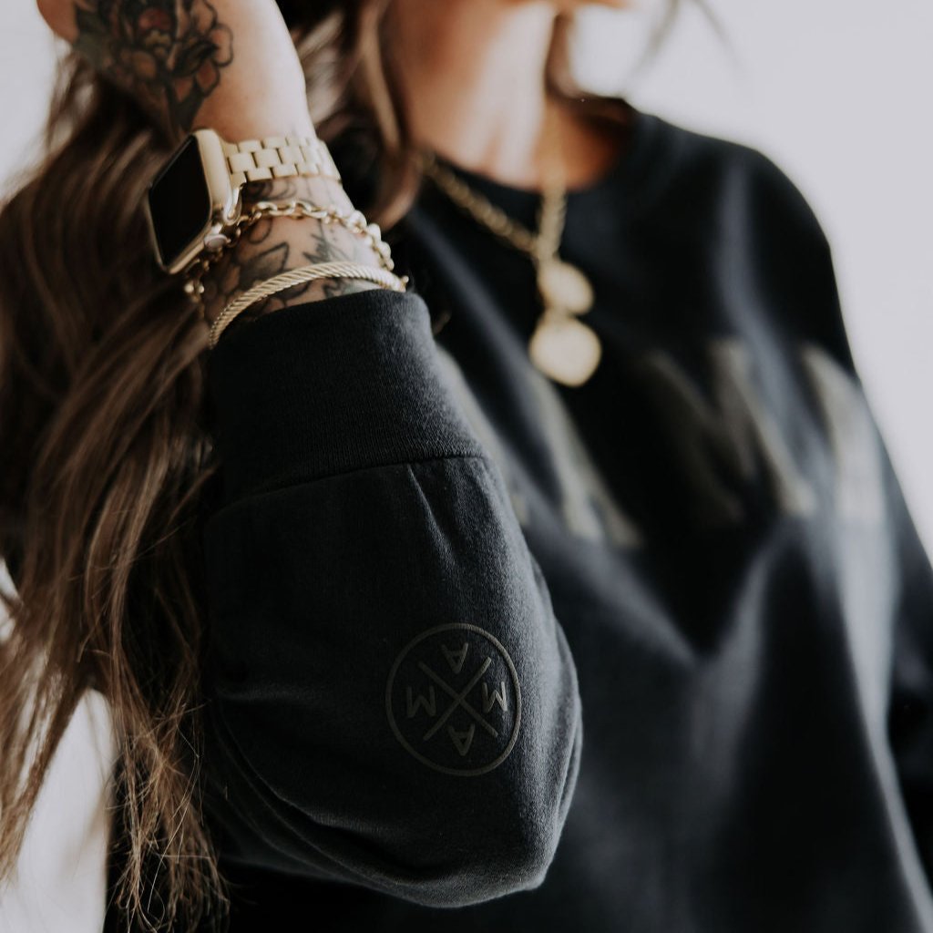 A person in a Mama X™ Black-out Crewneck, featuring a puff print circular logo on the sleeve. Their arm is tattooed, and they have long hair. They sport a smartwatch and a gold necklace with pendant. The bright and blurred background adds an intriguing aura to this ensemble from the Mama X™ Brand.