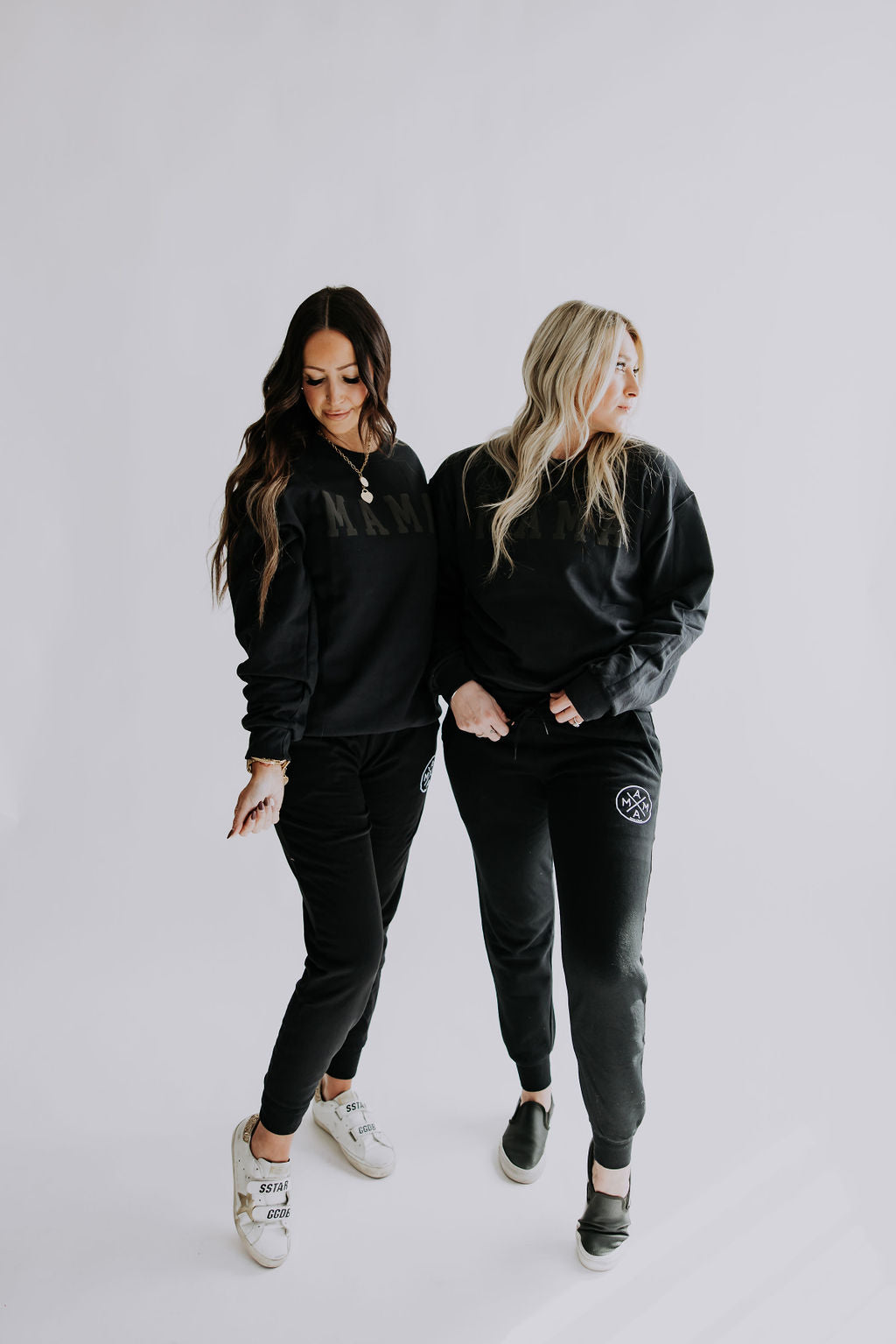 Two women in black matching outfits strike a pose against a plain backdrop. They are both dressed in classic fit sweatshirts paired with Mama X™ Joggers from Mama X™ Brand. One completes her look with sneakers, while the other opts for slip-on shoes. Both exude an easy-going everyday style, gazing in different directions with relaxed postures.