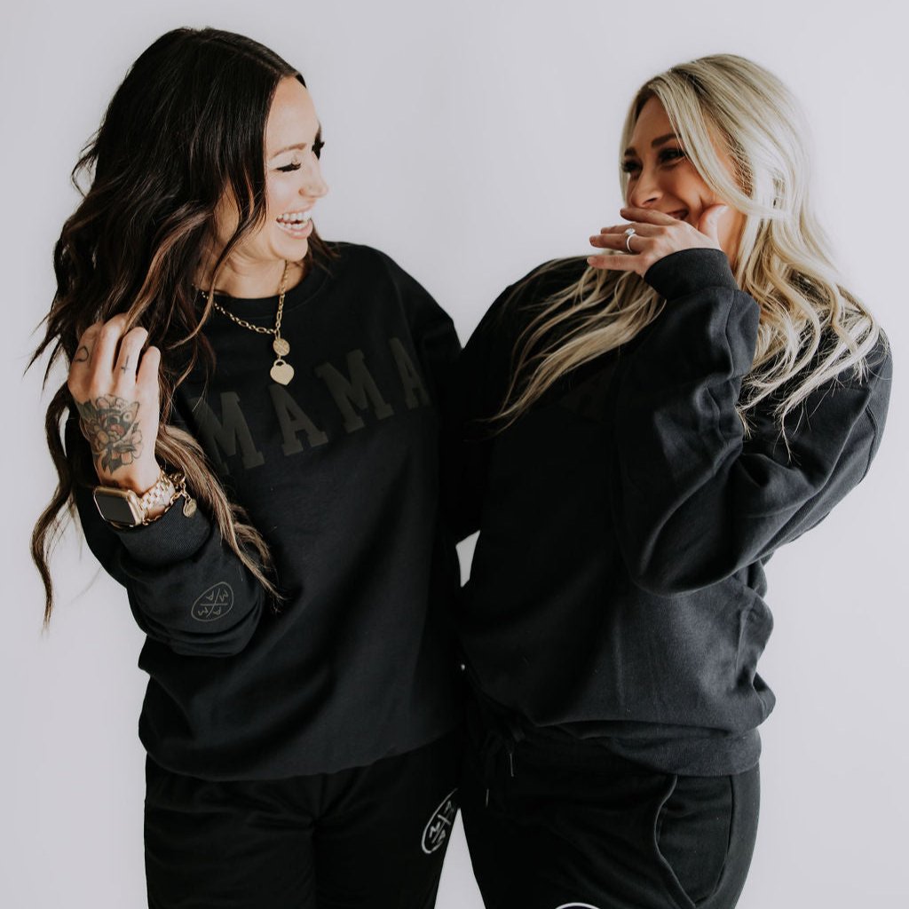 Two women in black Mama X™ Black-out Crewnecks stand closely together, smiling and laughing. One has long dark hair and a tattooed hand, while the other sports long blonde locks. The playful puff print from the Mama X™ Brand on their sweatshirts adds a fun touch against the plain white background.