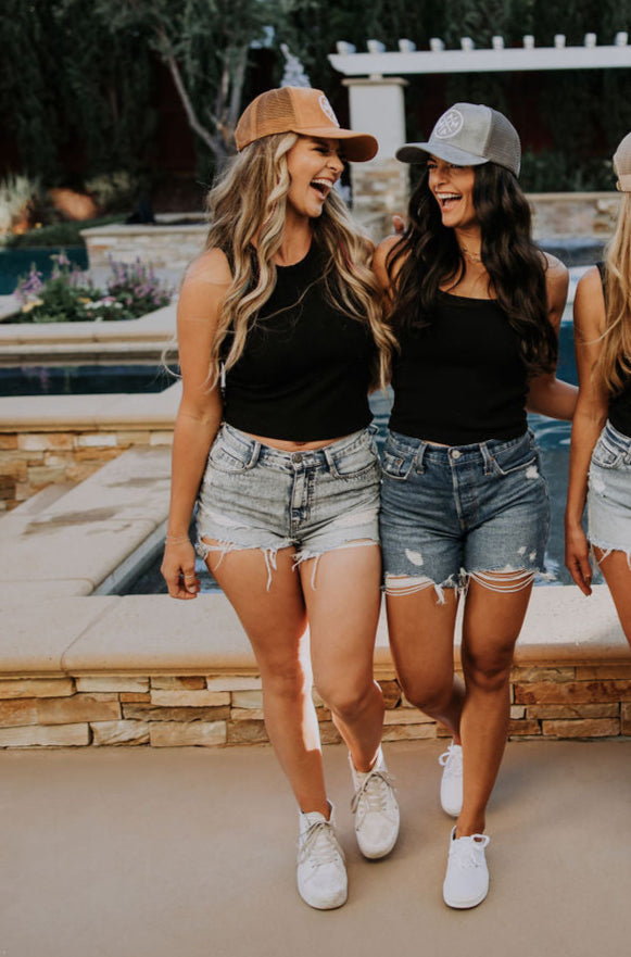Four women in matching black tops, denim shorts, and chic Mama X™ Trucker Hats in gray suede stand in a row, smiling at each other by a pool with tall trees in the background.