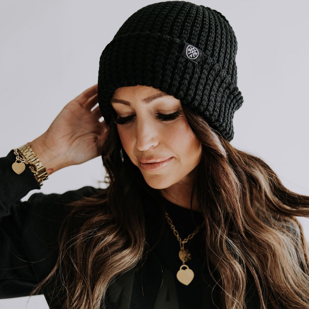 A person with long hair wears the Mama X™ Toasty Waffle Knit Beanie in black, coordinating it with a matching outfit. Their look is accessorized with a gold necklace and bracelet. They pose stylishly, one hand gently touching their beanie against a neutral background, perfectly embodying the essence of winter fashion.