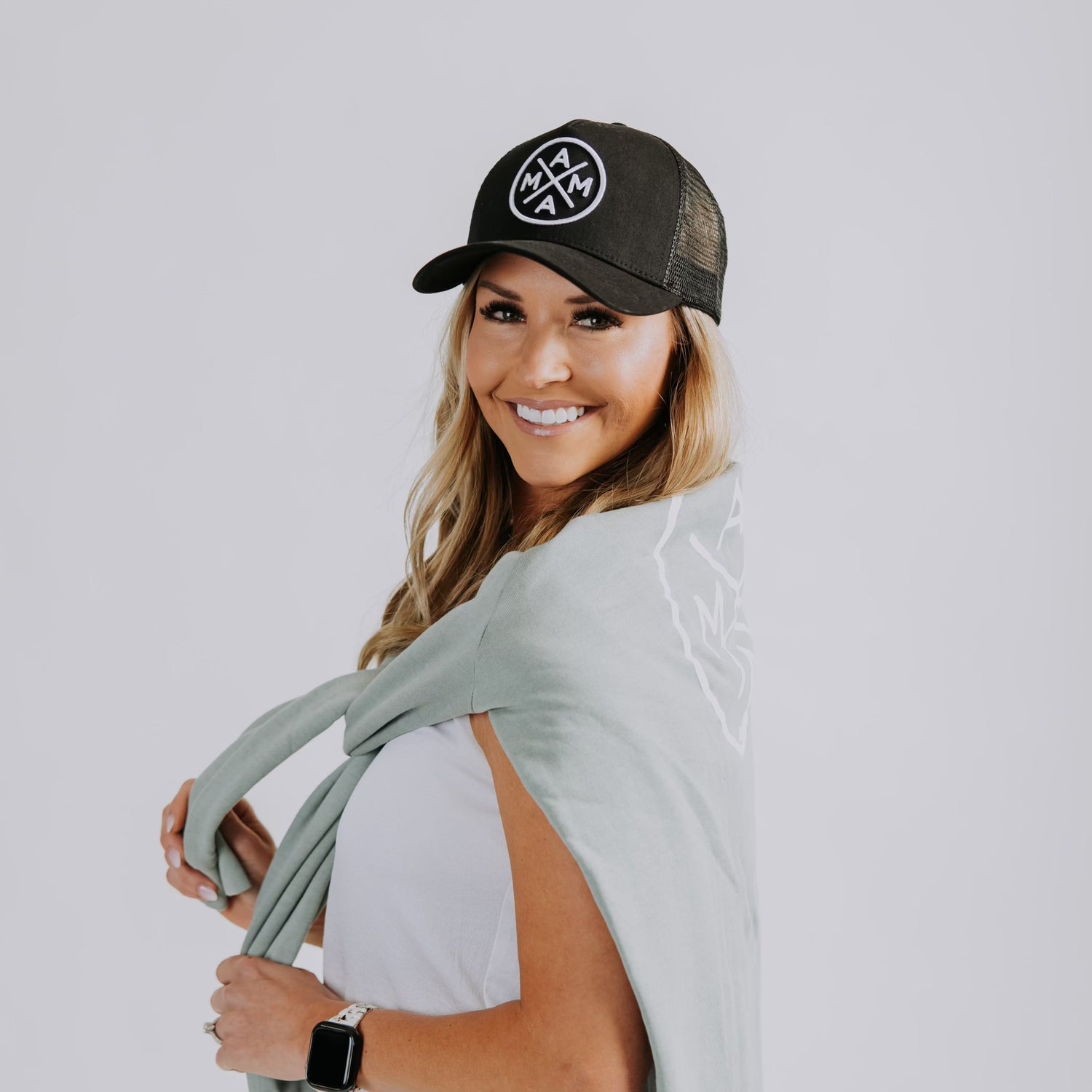 A woman smiles at the camera, wearing a Mama X™ Premium Canvas Trucker Hat in black with the MAMA X logo and a light green scarf draped over her shoulders. She is dressed in a white top and accessorized with a black smartwatch. The background is a plain light gray.