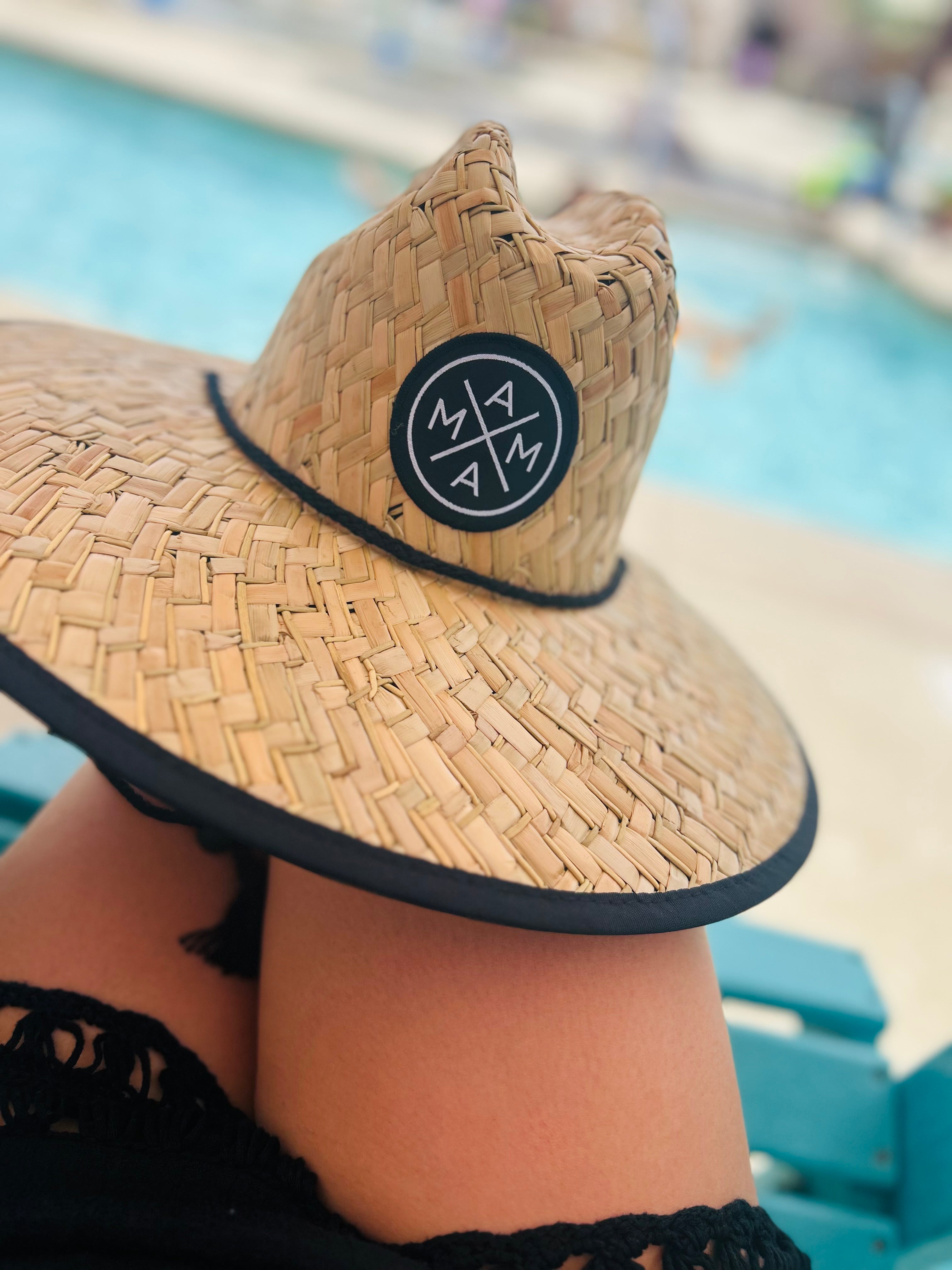 The Mama X™ Straw Hat by Mama X™ Brand provides stylish sun protection with its wide brim and signature round patch, seen resting on crossed legs poolside with a blurred water backdrop.