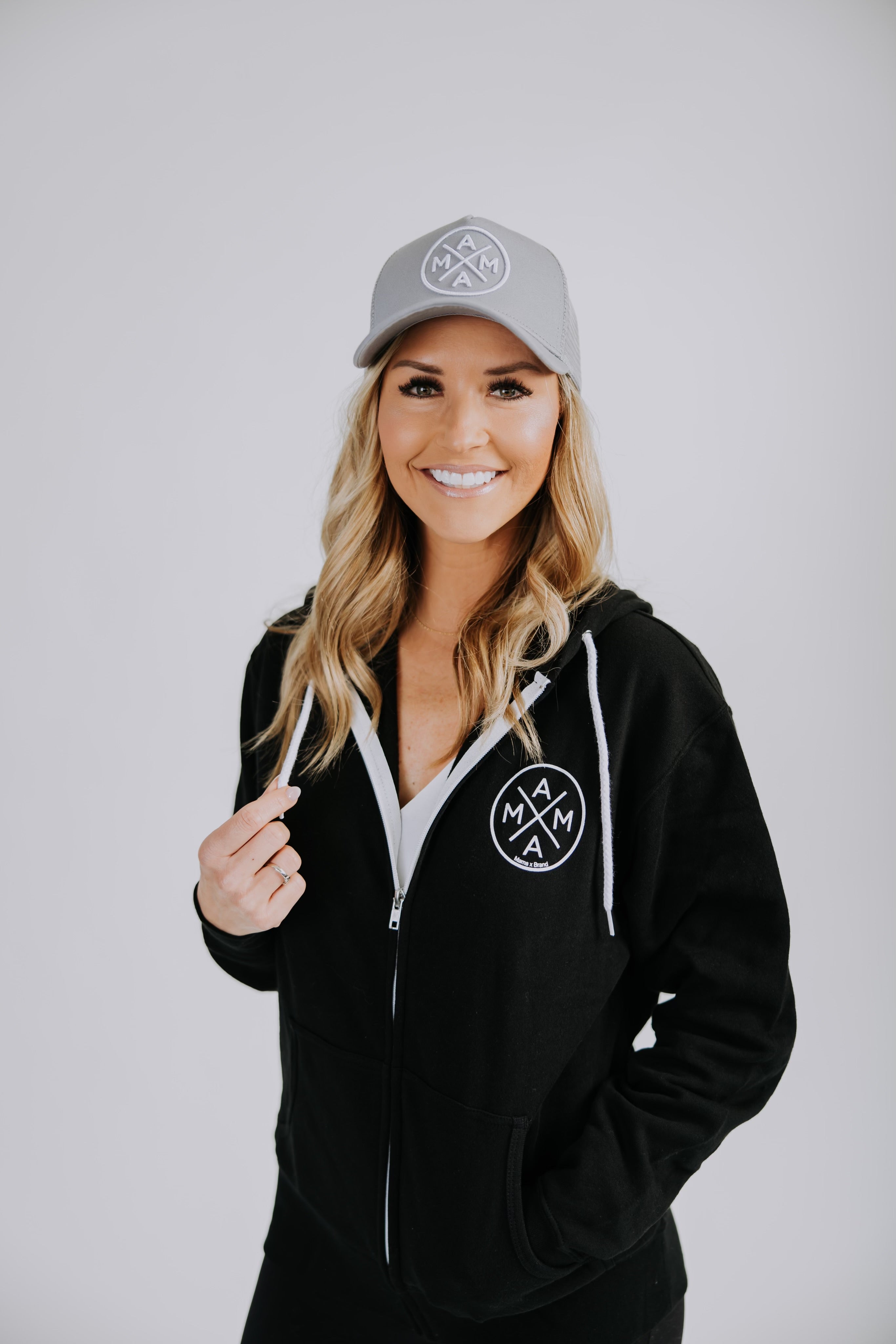 A smiling woman with long, wavy blonde hair wears a grey baseball cap and the Mama X™ Zip Up Sweatshirt - Black from Mama X™ Brand, both featuring a circular logo. She stands against a plain light grey background, touching the drawstring of the sweatshirt with her left hand.