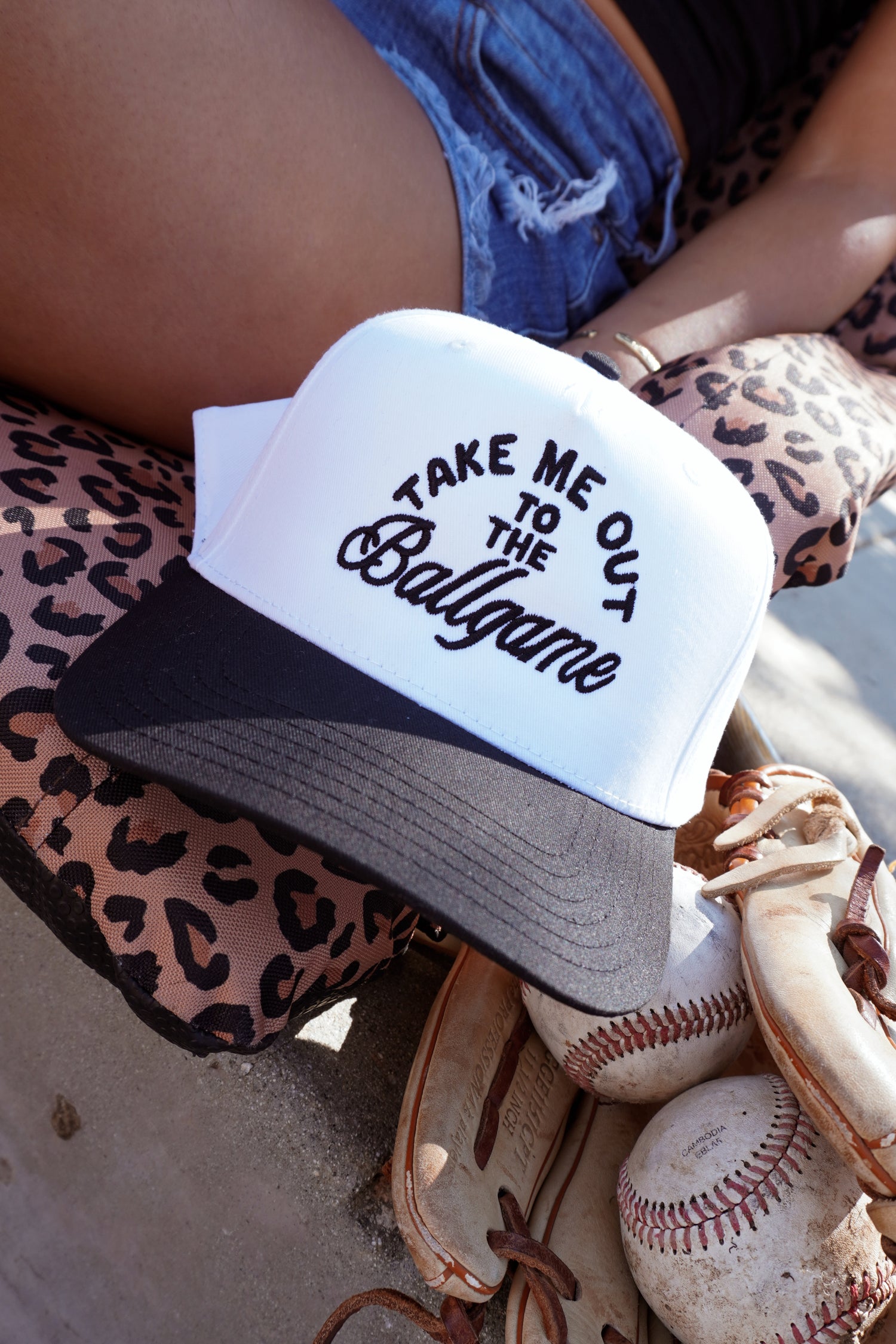 The Mama X™ Brand Ballgame Trucker, a white and black cap with "Take Me Out to the Ballgame," sits on a leopard print cushion beside a baseball glove and ball. A person in denim shorts is partly visible behind.