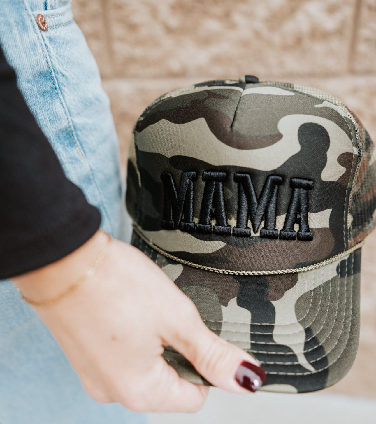 A person with dark red nail polish and a gold bracelet holds the Mama X™ Brand Black Camo Trucker hat, featuring "MAMA" embroidered in black, while wearing light blue jeans and a black top.