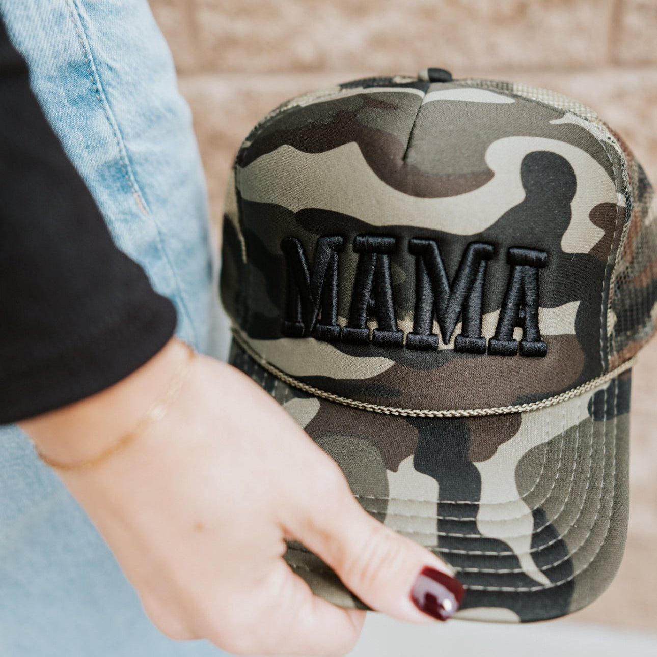 A person with dark red nail polish and a gold bracelet holds the Mama X™ Brand Black Camo Trucker hat, featuring "MAMA" embroidered in black, while wearing light blue jeans and a black top.