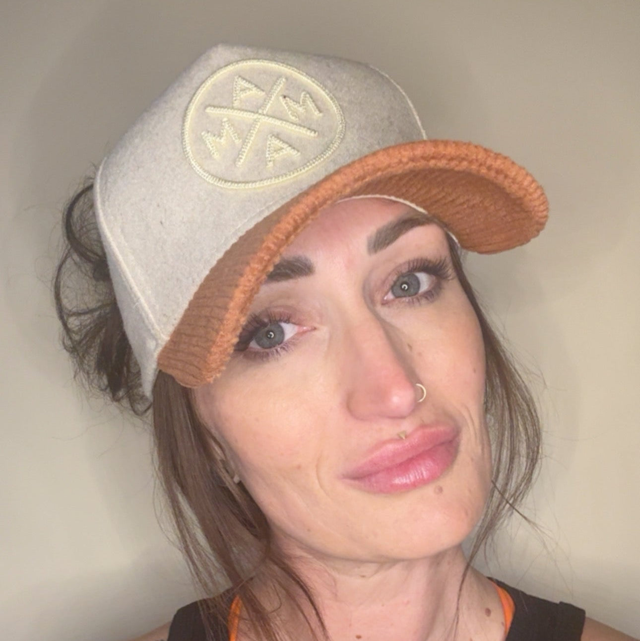 A woman with long brown hair and tattoos models Courtney’s Trucker hat by Mama X™ Brand—a beige and brown felt cap with a corduroy brim—paired with a black sleeveless top, nose ring, and subtle makeup against a light background.