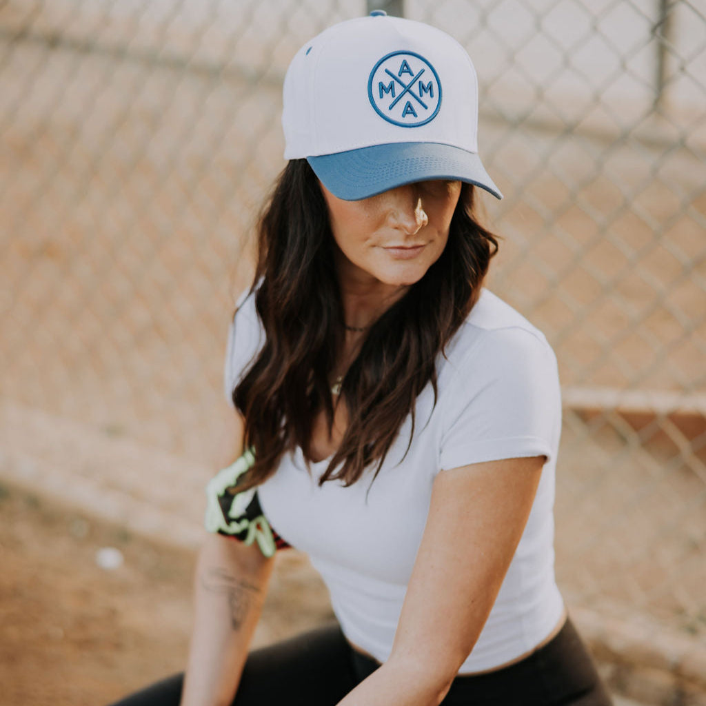 A person with long hair kneels by a fence wearing a Blue Mama X™ Baseball Cap featuring the intertwined "A" and "M" logo. They&