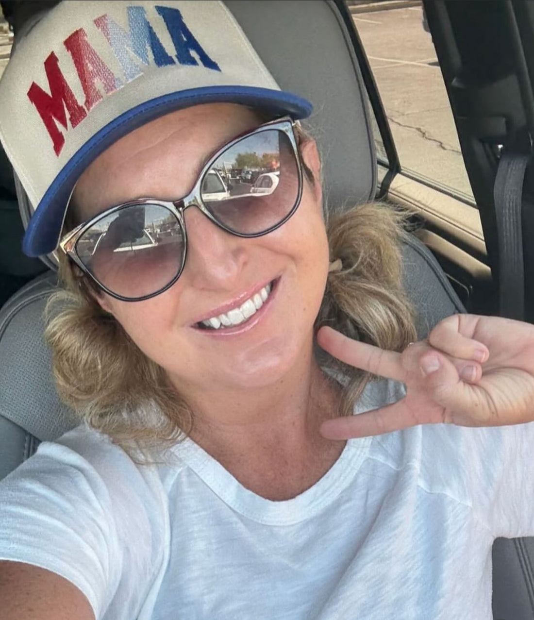 A woman in sunglasses and a white shirt smiles, flashing a peace sign in her car. She wears the Red, White &amp; Blue Mama Hat by Mama X™ Brand, with blonde hair peeking from beneath its bold embroidered design.