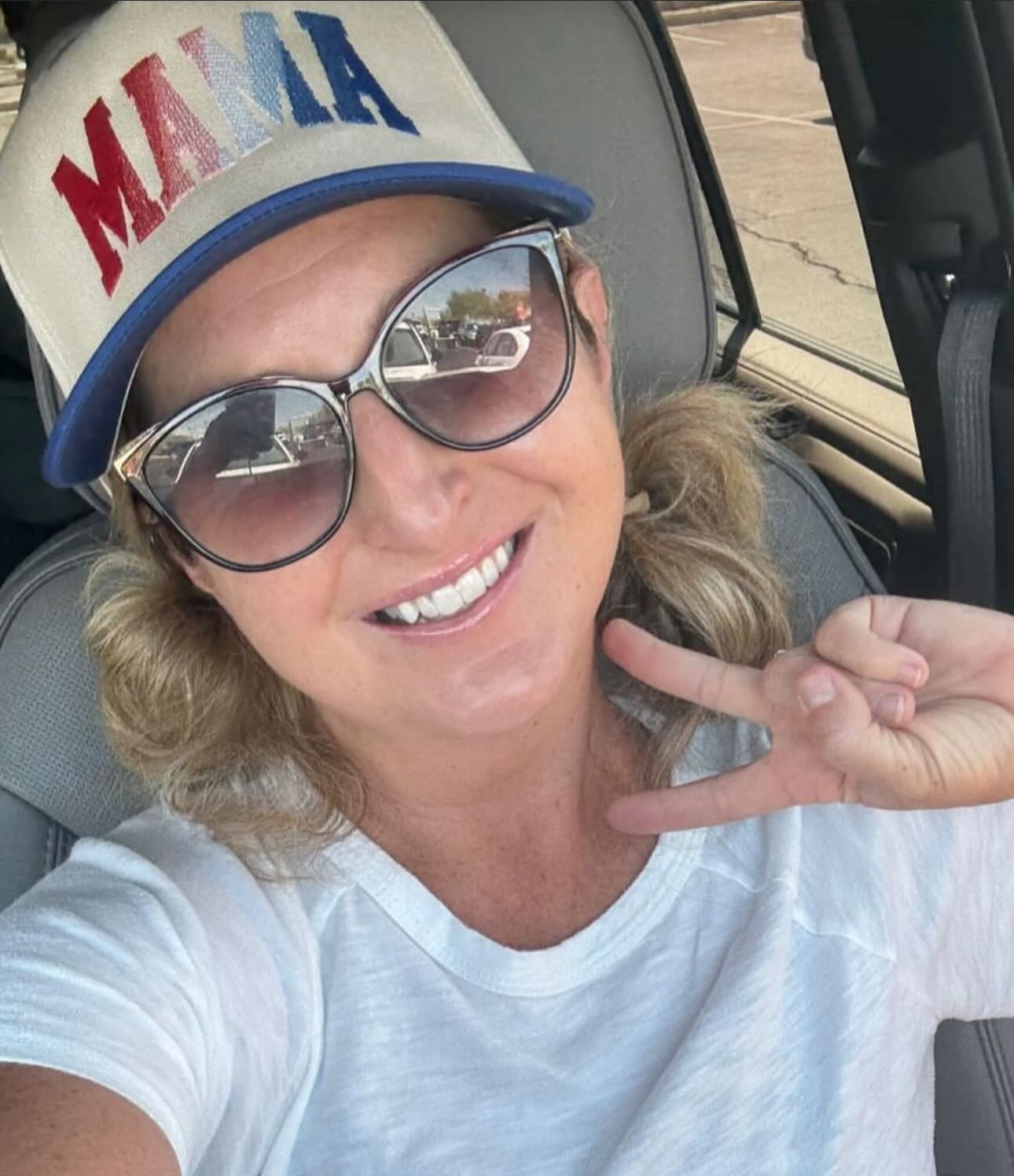 A woman in sunglasses and a white shirt smiles, flashing a peace sign in her car. She wears the Red, White & Blue Mama Hat by Mama X™ Brand, with blonde hair peeking from beneath its bold embroidered design.