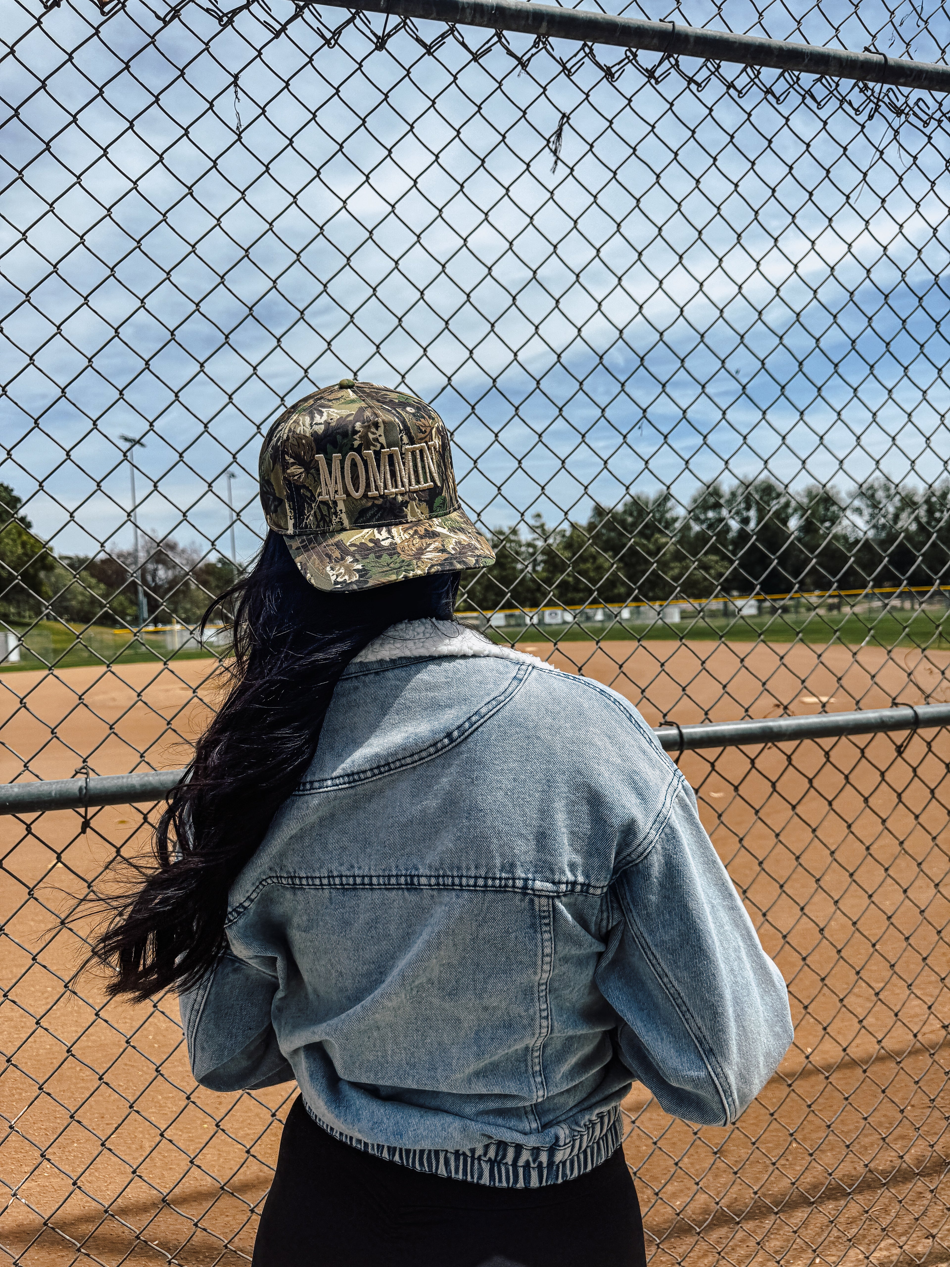A woman with long dark hair, in a denim jacket and a Camo Mommin’ cap by Mama X™ Brand, stands behind a chain-link fence overlooking a baseball field on a sunny day.