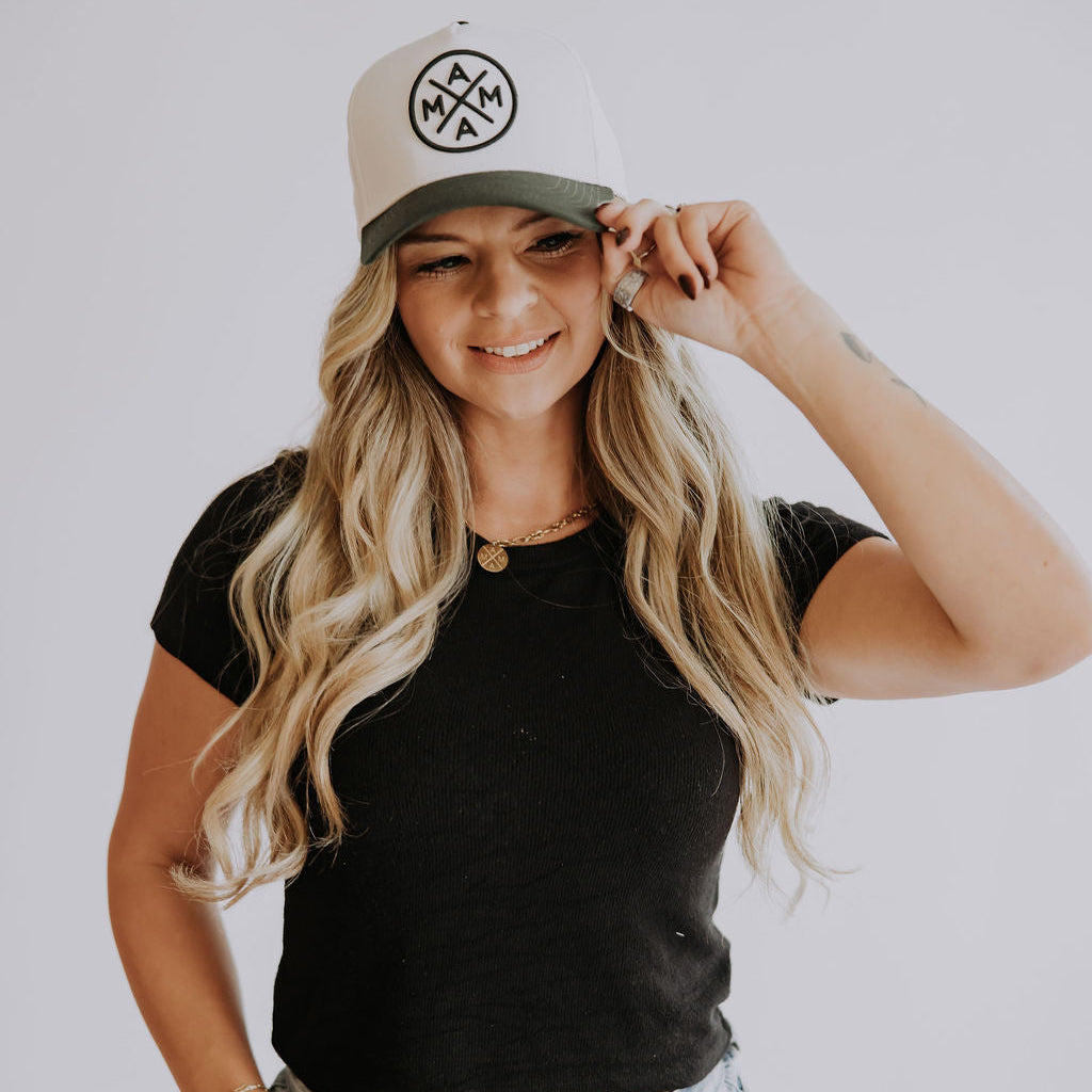 A woman with long, wavy blonde hair, wearing a black crop top, light blue jeans, and a dark green Mama X™ Baseball Cap by Mama X™ Brand, smiles as she adjusts her hat with one hand against a plain white background.