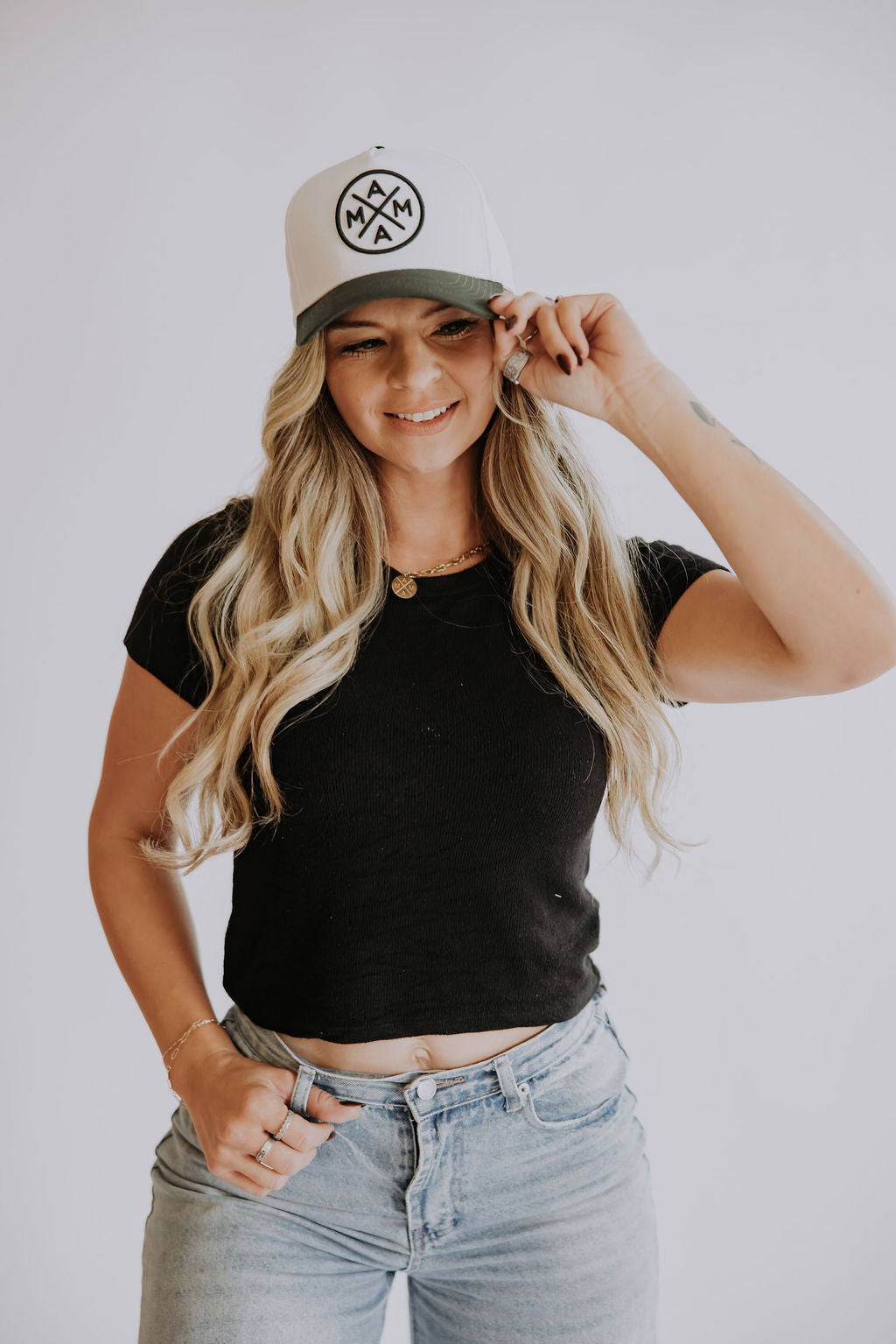 A woman with long, wavy blonde hair, wearing a black crop top, light blue jeans, and a dark green Mama X™ Baseball Cap by Mama X™ Brand, smiles as she adjusts her hat with one hand against a plain white background.