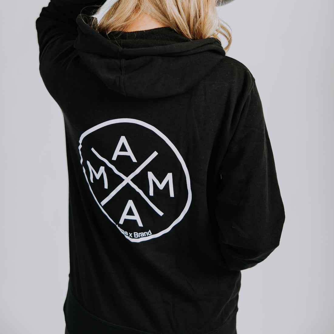 A person with long blonde hair, shown from behind, wears a Mama X™ Brand fleece zip-up hoodie with a large "MAMA" logo on the back and a light gray mesh cap, touching the cap with one hand.