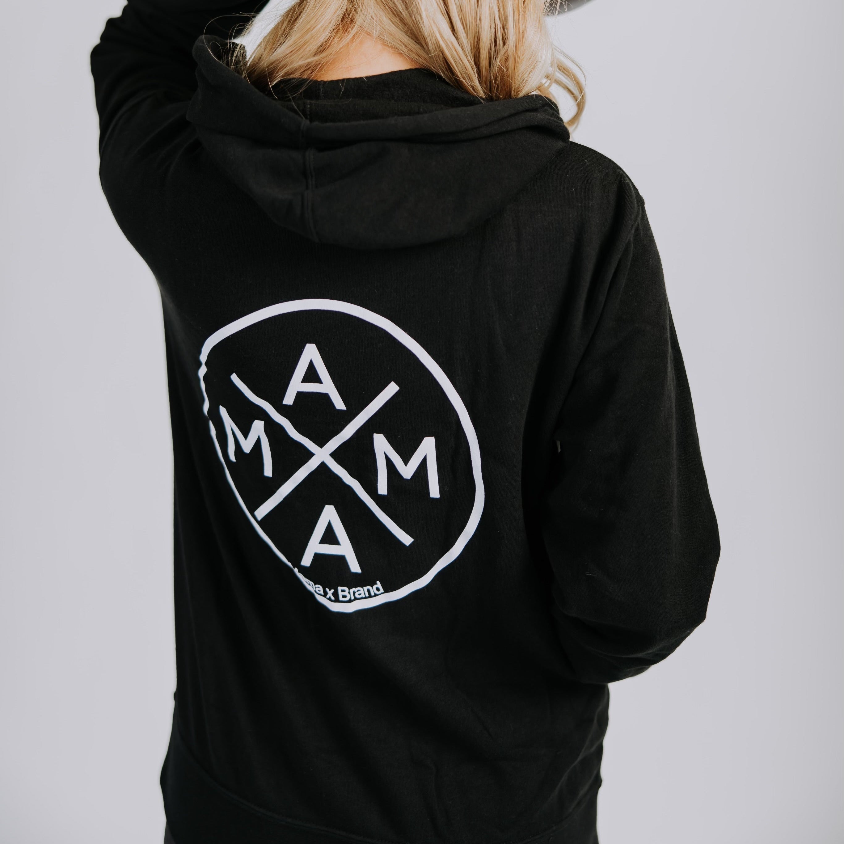 A person with long blonde hair, shown from behind, wears a Mama X™ Brand fleece zip-up hoodie with a large "MAMA" logo on the back and a light gray mesh cap, touching the cap with one hand.