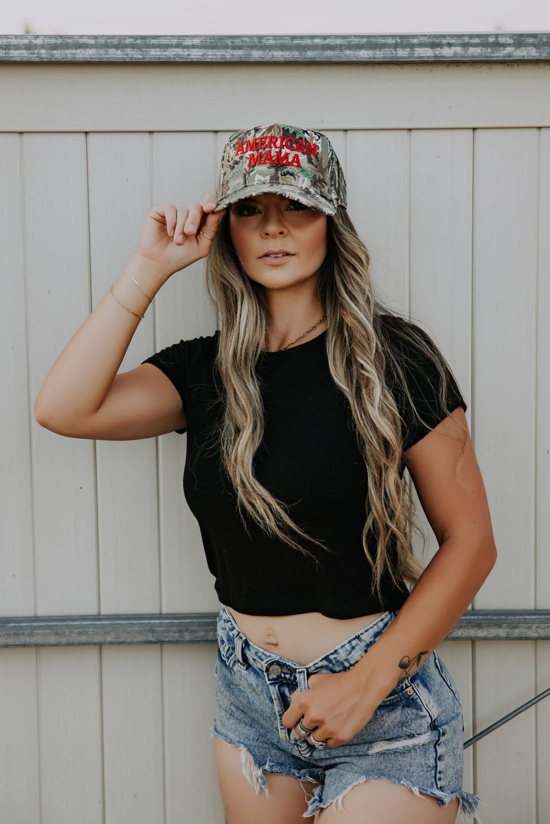 A woman with long, wavy hair wears the American Mama Hat by Mama X™ Brand— a cotton twill camo cap with bold red lettering—paired with a black crop top and denim shorts, standing in front of a white paneled fence.