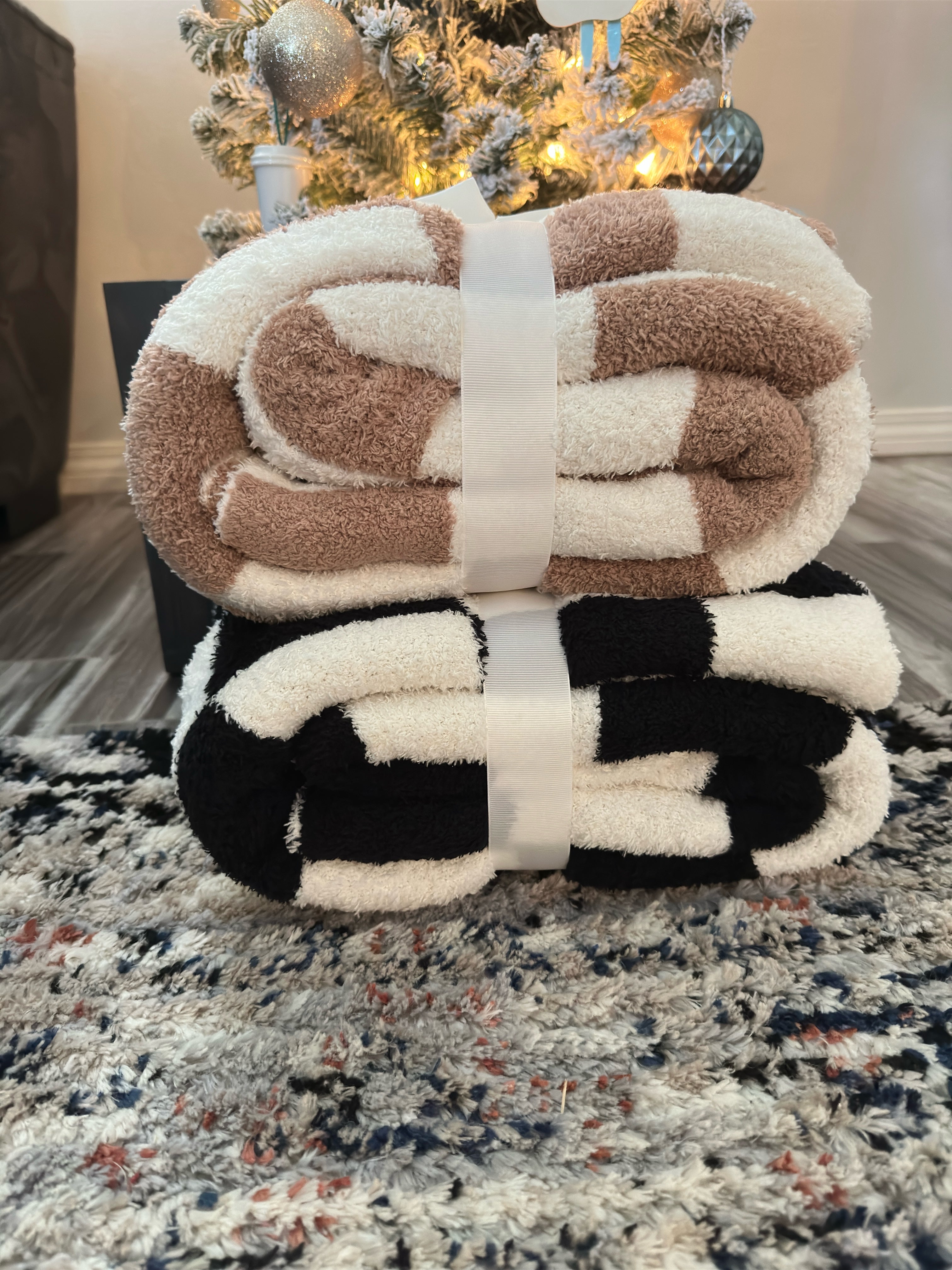 Two folded, striped blankets from the Mama X™ Brand are cozily stacked under a Christmas tree. The top blanket is Mama X Checkered Blanket in soft beige and white, while the bottom one features a stylish black and white design. They rest on a textured, multicolored rug with holiday decorations in the background.