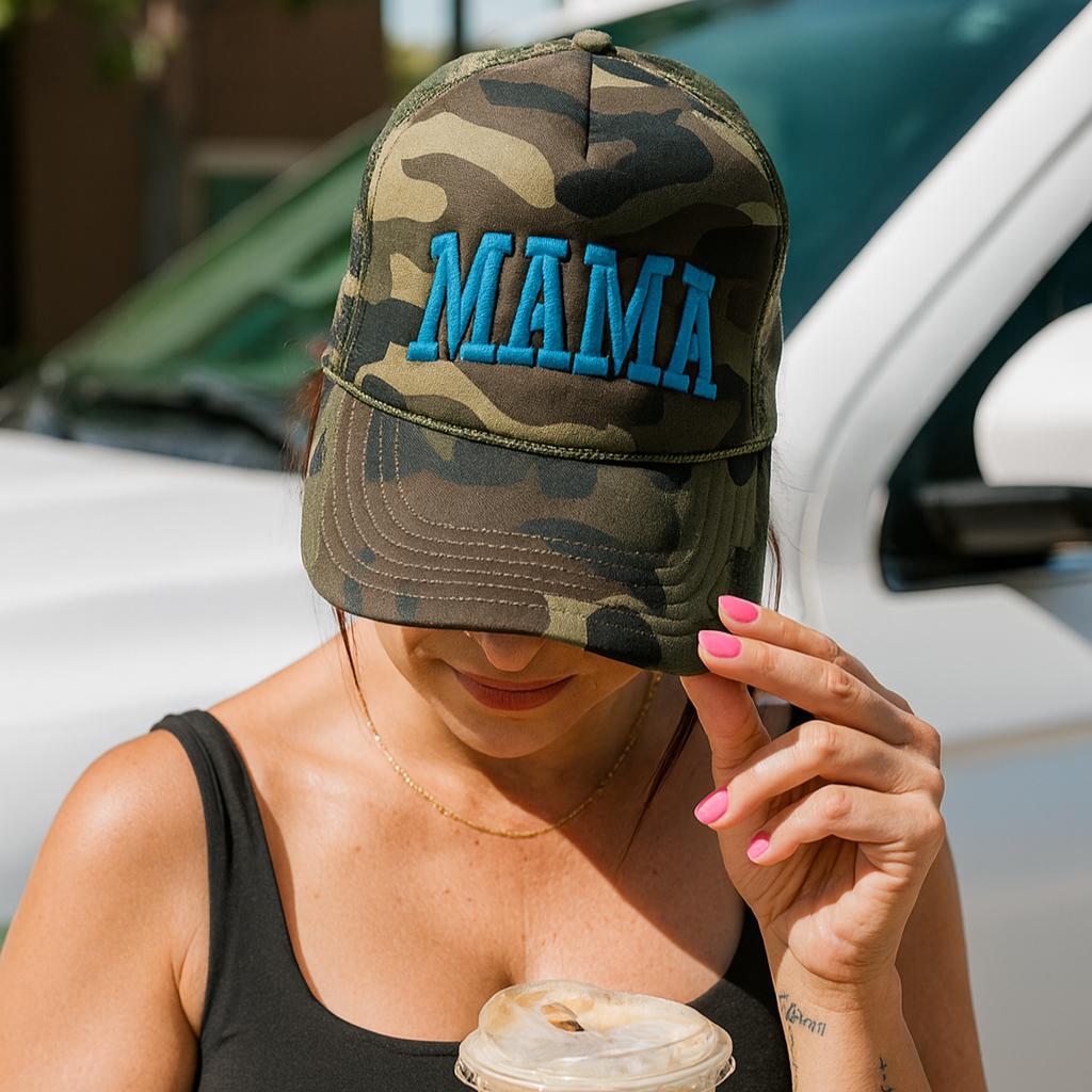 A woman in a black tank top holds an iced coffee, showing off pink nail polish and wearing the Mama X™ Brand Camo/Blue Mama Foam Hat, standing by a white vehicle.