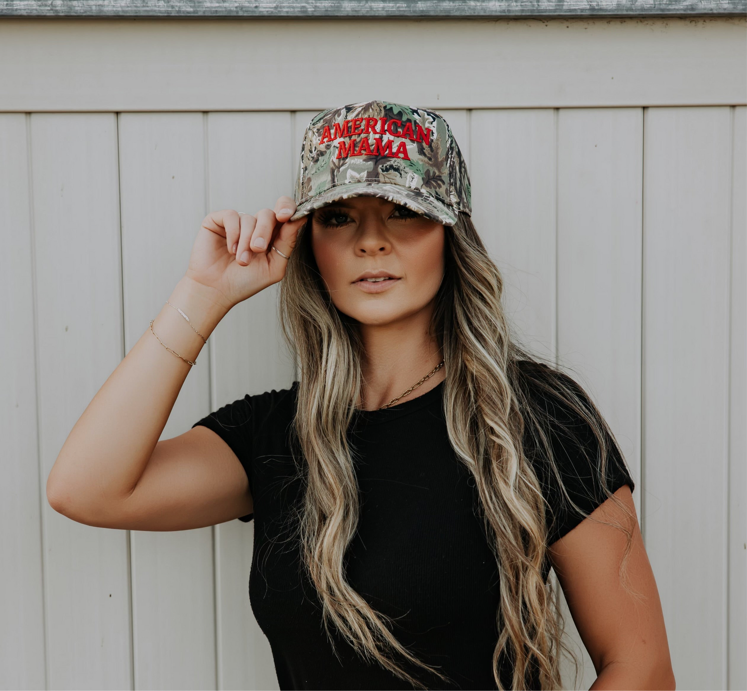 A woman with long, wavy hair wears the Mama X™ Brand American Mama Hat—a camo cap with "AMERICAN MAMA" in red—paired with a black T-shirt, standing before a white wooden fence and holding the brim of her hat.