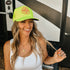 A smiling woman with long wavy hair wears the Neon Yellow Mama X Trucker by Mama X™ Brand and a white tank top, sitting outdoors in front of an RV. She holds a ring with blue and red flowers and wears a gold necklace.