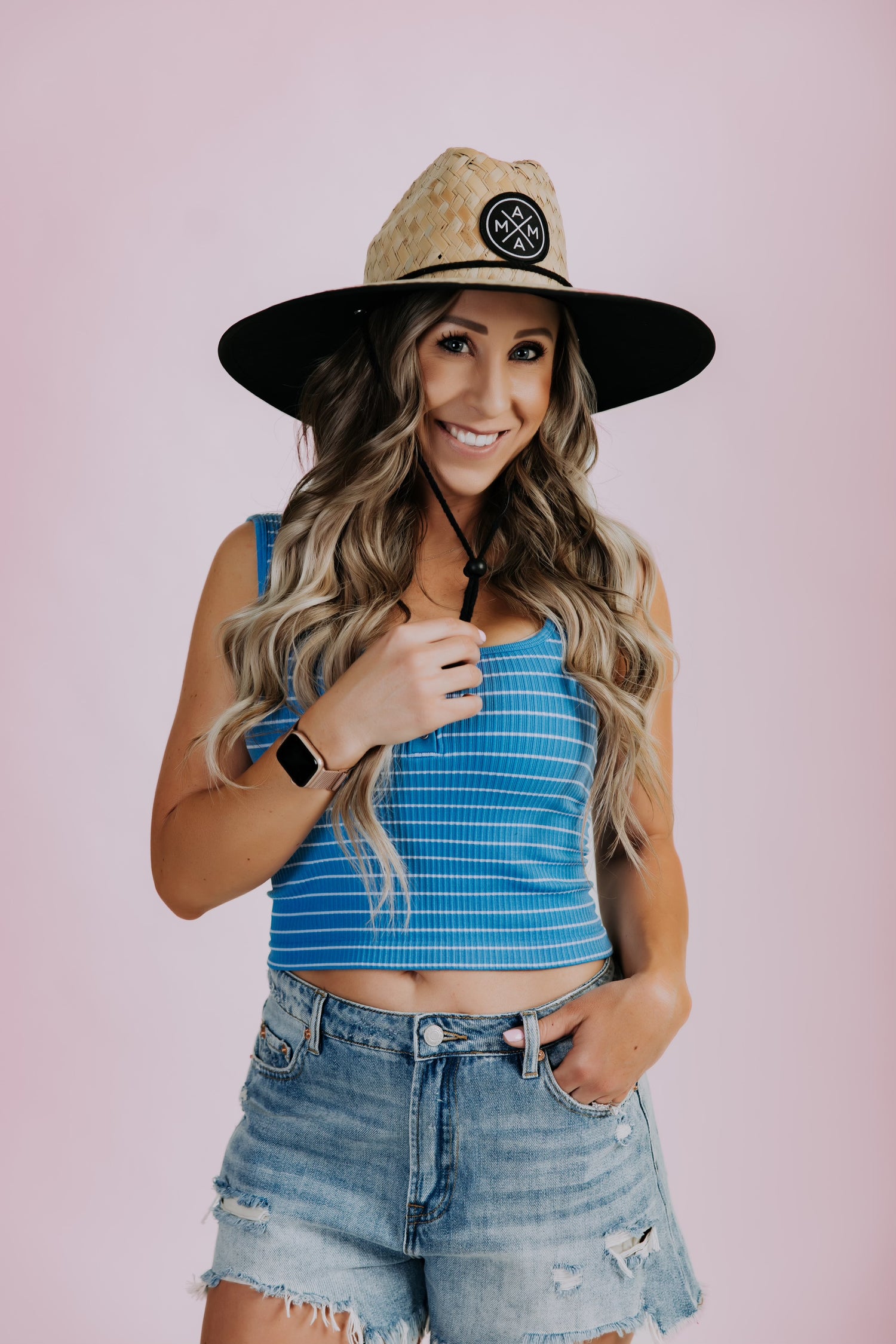Against a pink background, a woman in a blue cropped tank and ripped denim shorts poses with confidence. Her long wavy hair flows beneath the Mama X™ Brand&