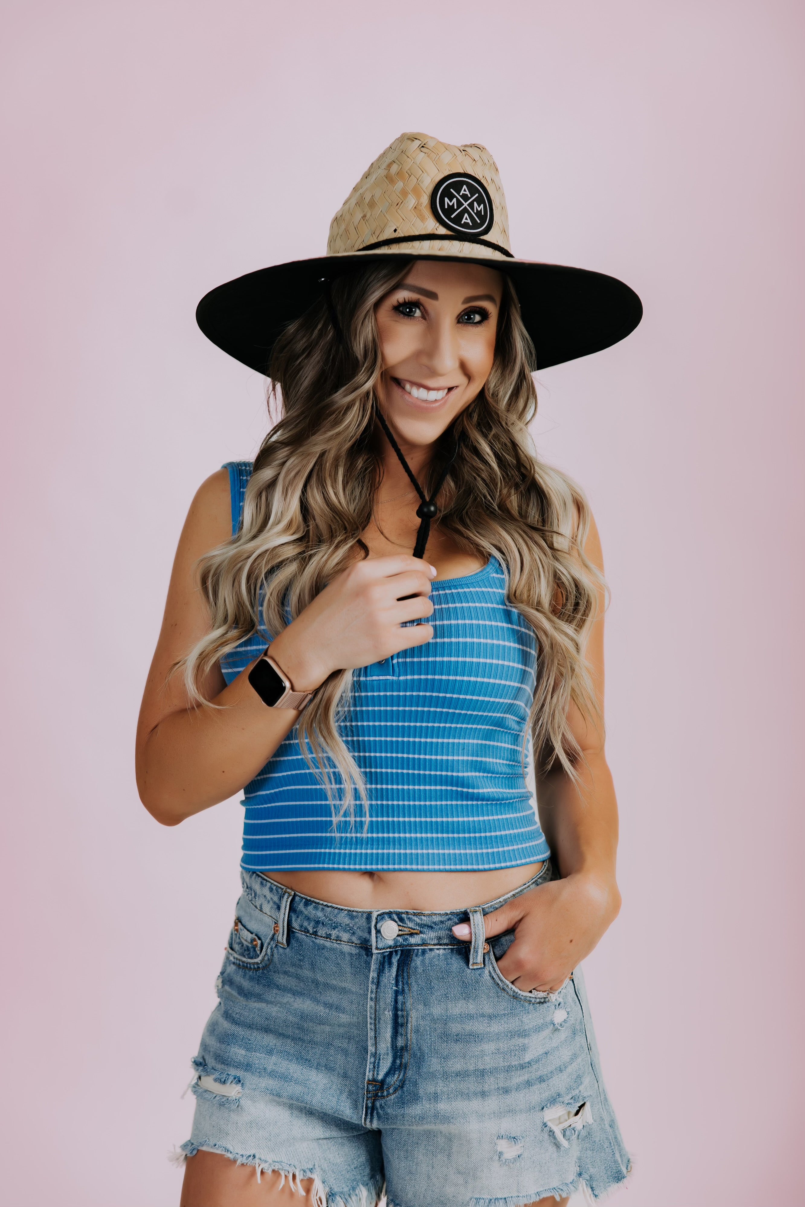 Against a pink background, a woman in a blue cropped tank and ripped denim shorts poses with confidence. Her long wavy hair flows beneath the Mama X™ Brand&