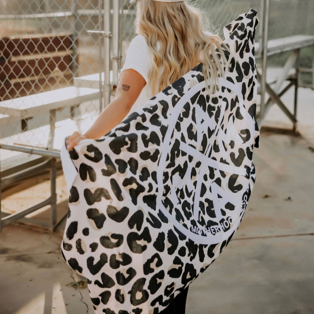 A person with long blonde hair, in a white shirt and hat, stands by metal bleachers at an outdoor sports facility holding the limited-edition "Motherhood Is So Fetch Towel" from Mama X™ Brand, featuring a leopard-print and white peace symbol.