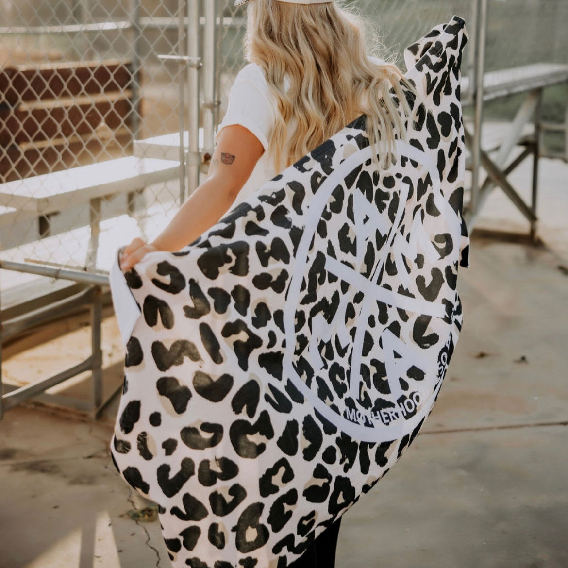 A person with long blonde hair, in a white shirt and hat, stands by metal bleachers at an outdoor sports facility holding the limited-edition "Motherhood Is So Fetch Towel" from Mama X™ Brand, featuring a leopard-print and white peace symbol.