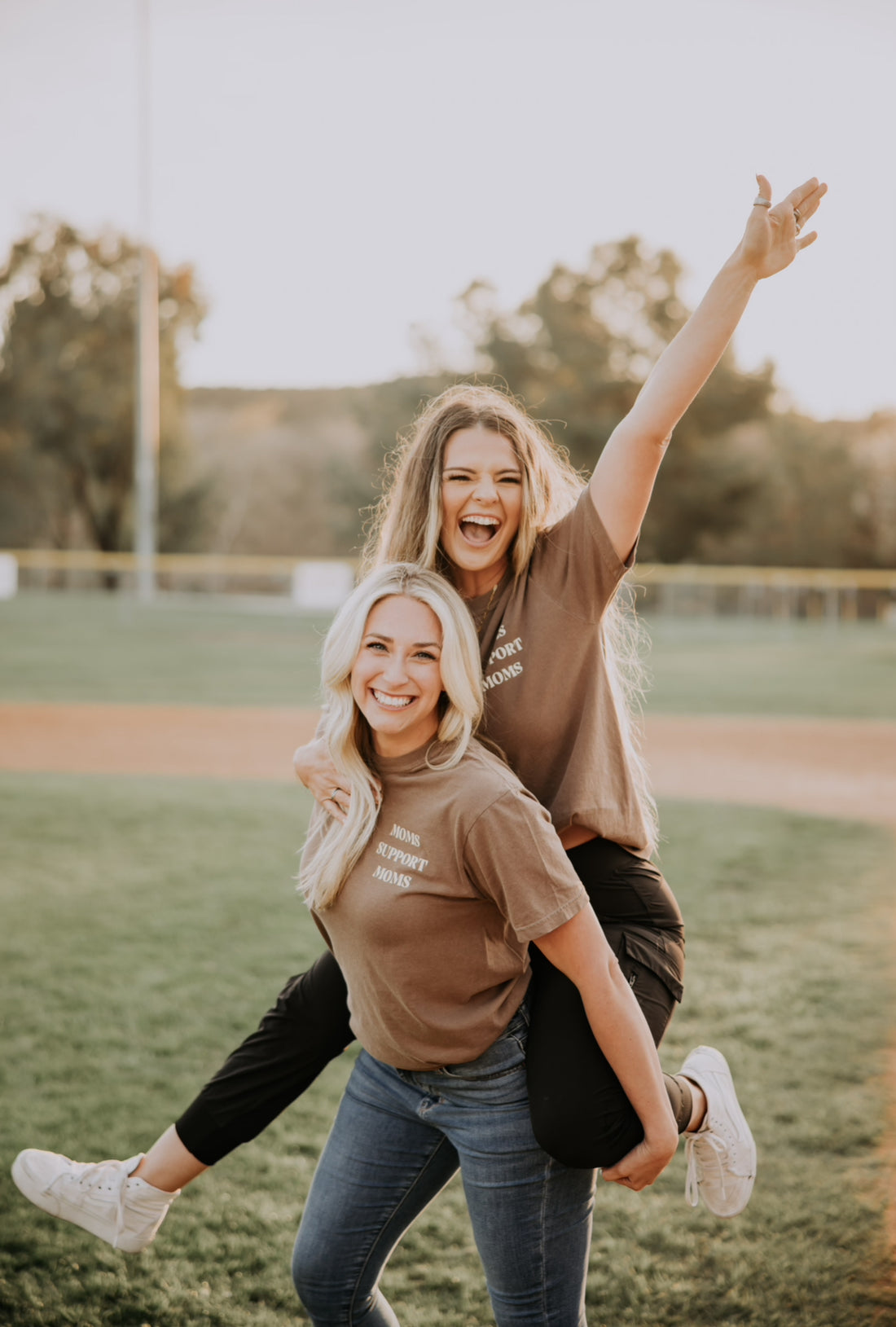 Two people are outside on a grass field, one giving the other a piggyback ride. Both wear Mama X™ Brand&