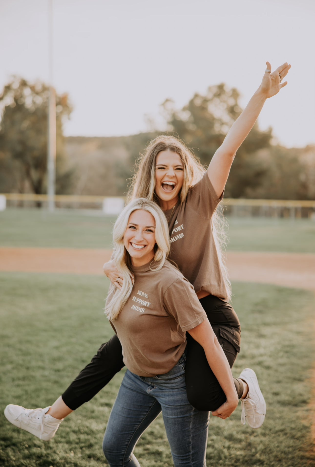 Two people are outside on a grass field, one giving the other a piggyback ride. Both wear Mama X™ Brand&