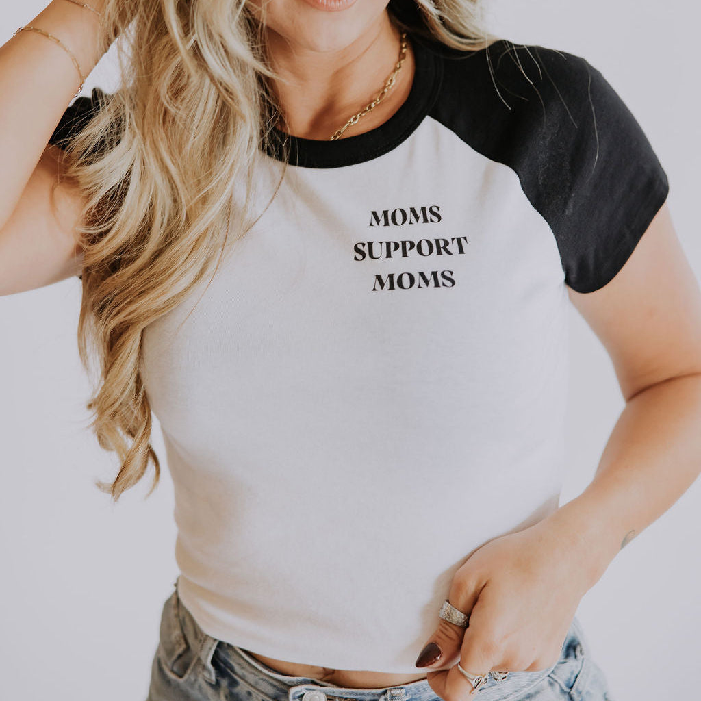 A woman with long, wavy blonde hair wears the Mama X™ Brand Moms Support Moms raglan baby tee, smiling gently with one hand in her hair against a plain light background.