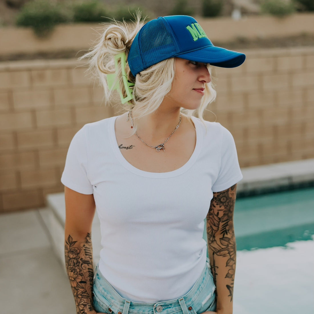 A tattooed woman by a pool wears the Mama X™ Brand Neon Blue/Green Foam Hat, paired with a white t-shirt and denim shorts. She stands near a brick wall and mountains, looking to the side with her hands in her pockets.