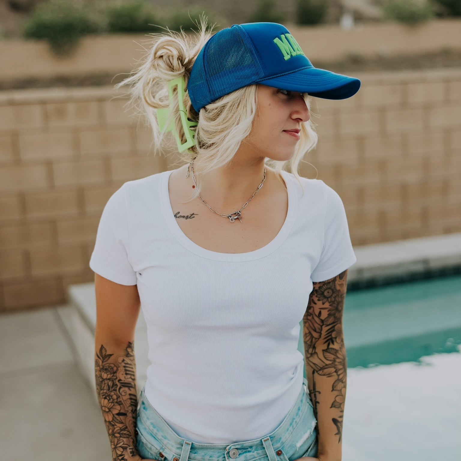 A tattooed woman by a pool wears the Mama X™ Brand Neon Blue/Green Foam Hat, paired with a white t-shirt and denim shorts. She stands near a brick wall and mountains, looking to the side with her hands in her pockets.