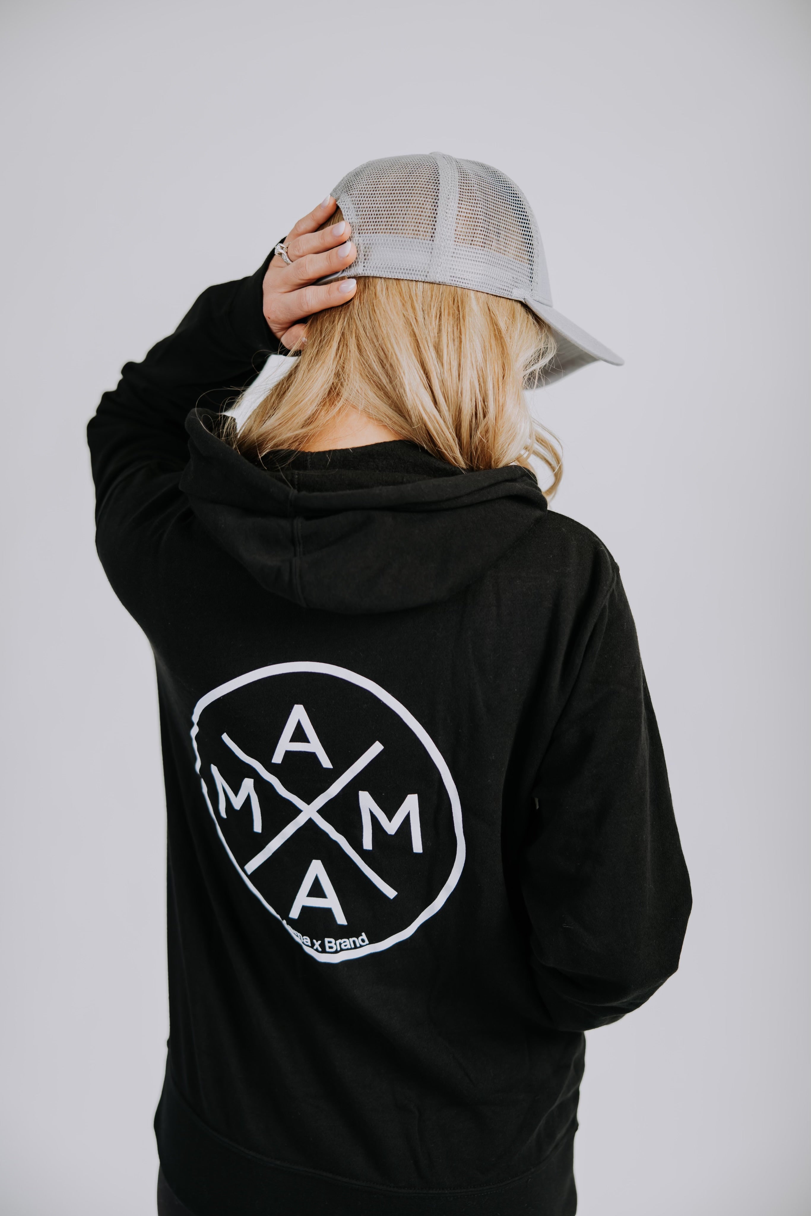 A person with long blonde hair is wearing the Mama X™ Zip Up Sweatshirt - Black and a grey baseball cap. The back of the hoodie, from the Mama X™ Brand and made from premium ring-spun cotton, features a circular logo with the letters 