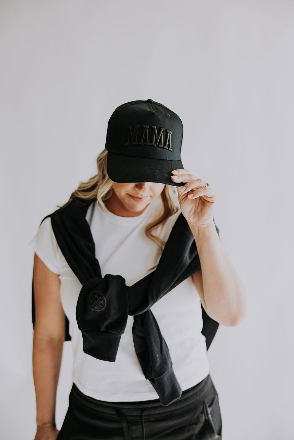 A woman in a white tee and black pants wears the Mama X™ Brand Mama Black on Black Trucker Hat with bold 3D embroidery, holding its brim and draping a black sweatshirt over her shoulders against a plain white background.