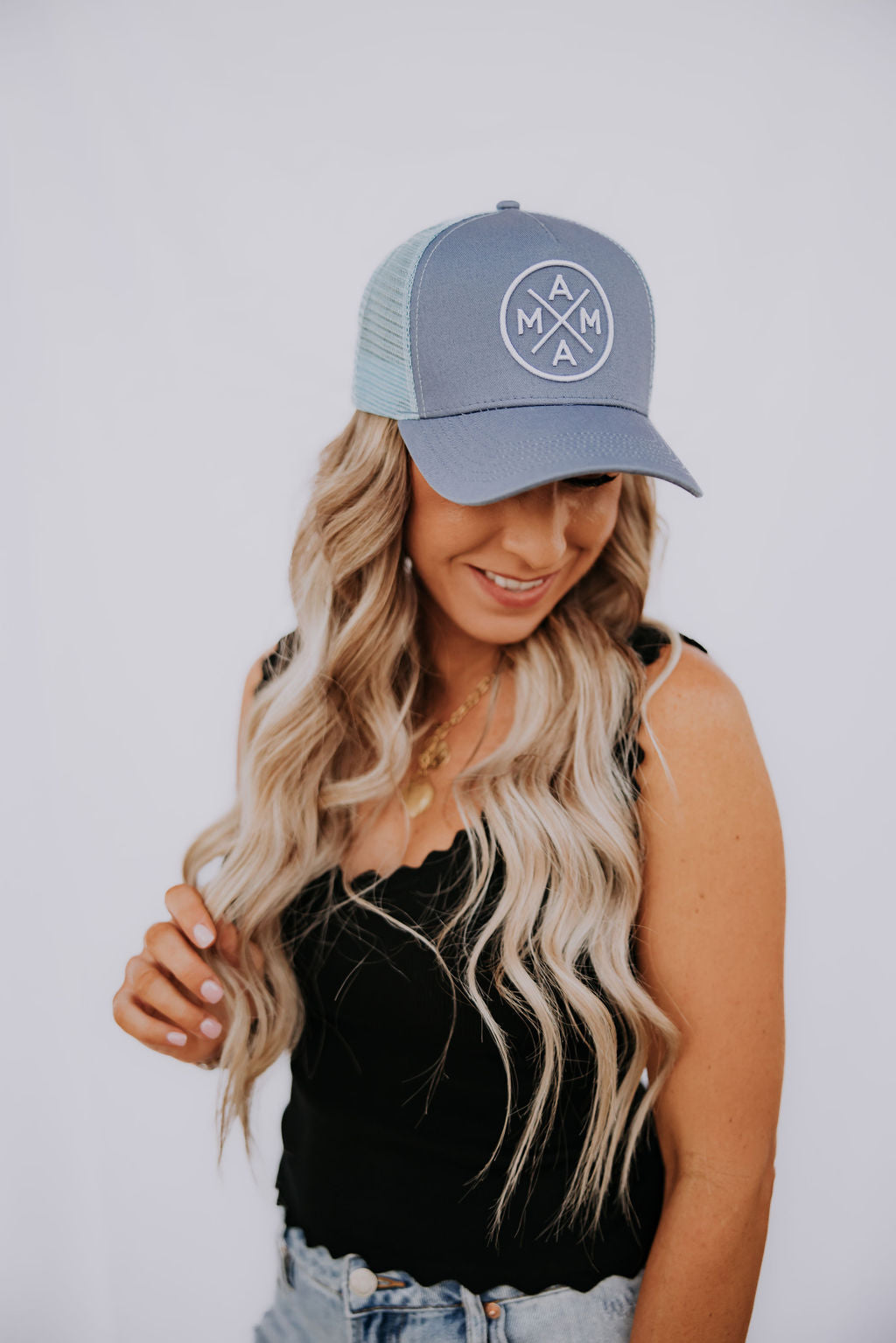 A woman with long, wavy blonde hair smiles and looks down against a white background. She wears a Mama X™ Brand Blue Mama X Trucker hat, a black tank top, and light blue jeans.
