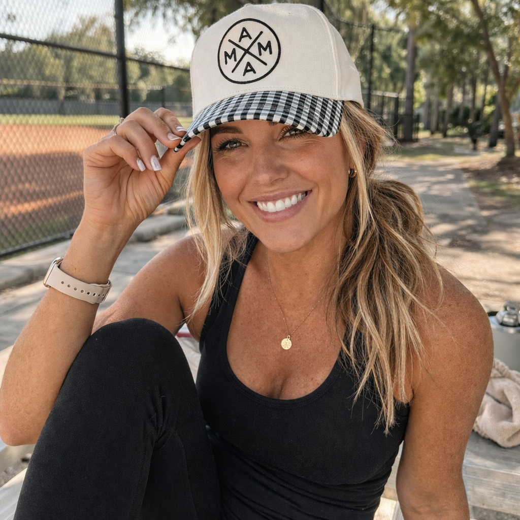 A woman in a Black Gingham Mama X Trucker by Mama X™ Brand and a black tank top smiles on a bench near the baseball field. With her ponytail and gold necklace, she embraces mom life and sunny days—her go-to everyday style.